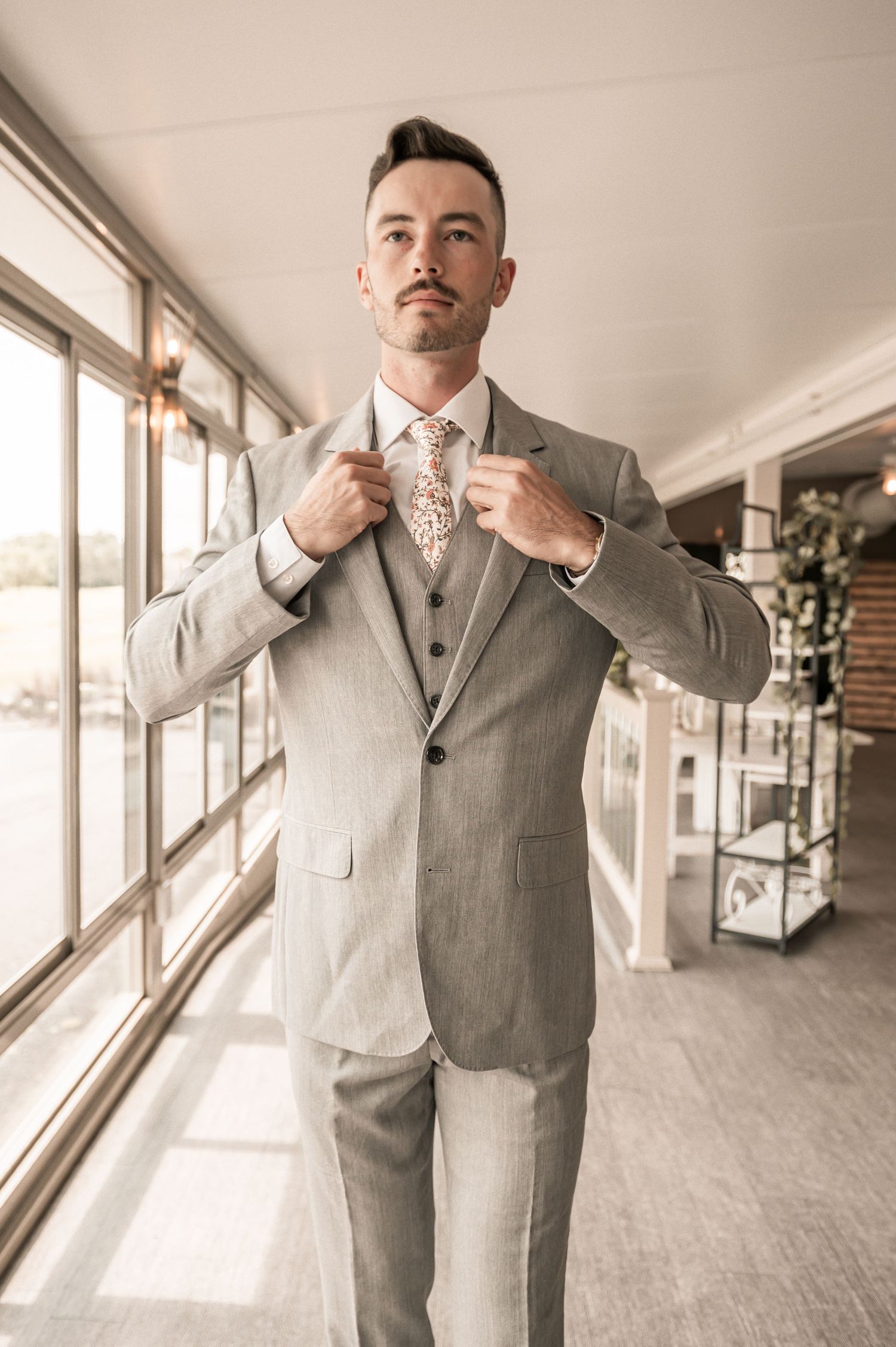 A person wearing a light gray three-piece suit with a patterned tie adjusts their jacket in a sunlit hallway.