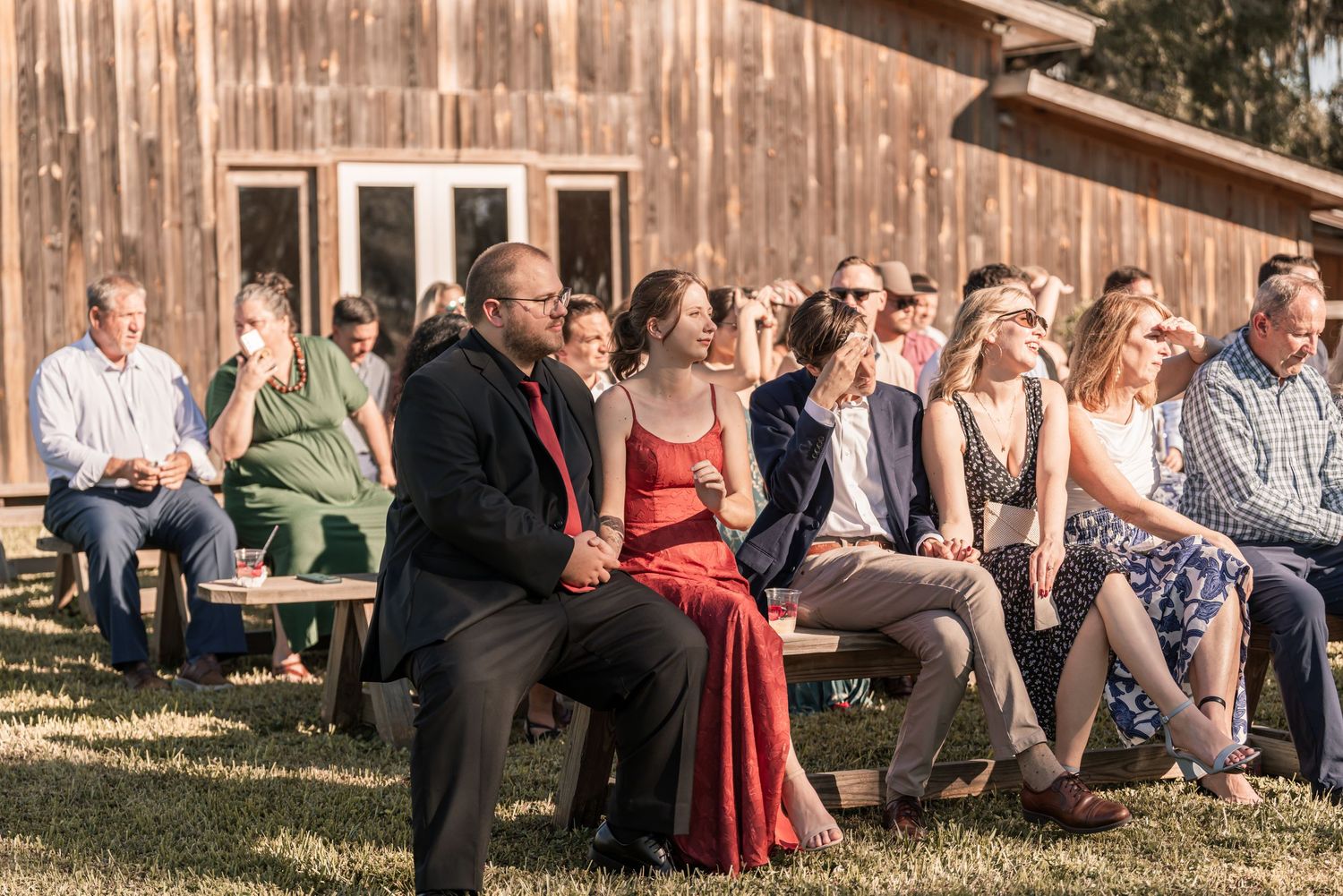 Wedding guests seated on wooden benches outdoors during a rustic ceremony at sunset.