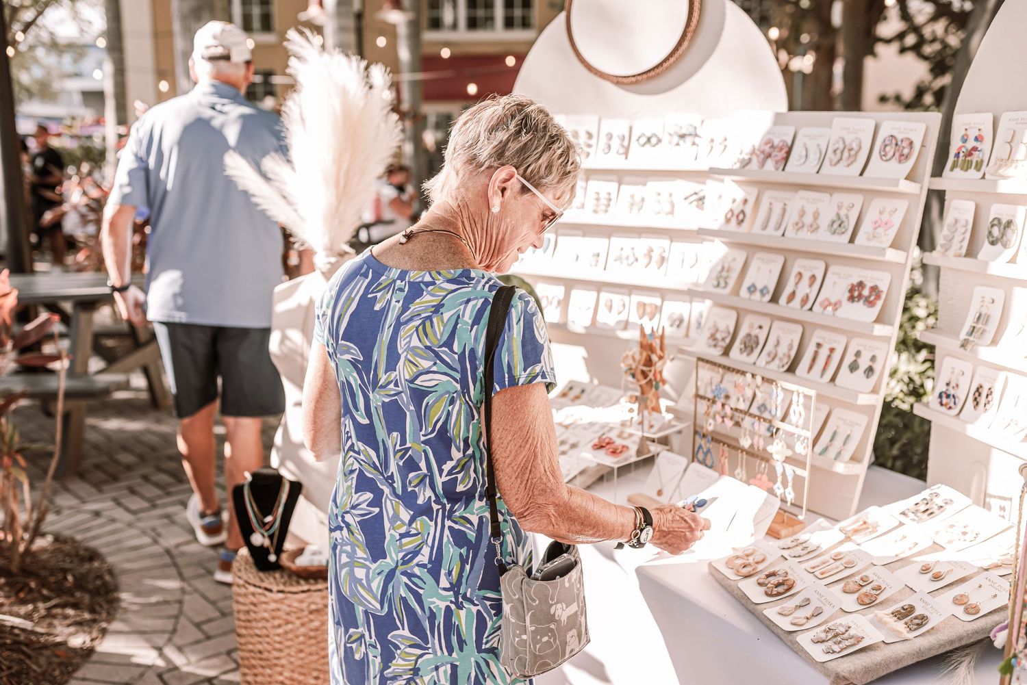 A shopper in a blue patterned dress browses jewelry at an outdoor market display.