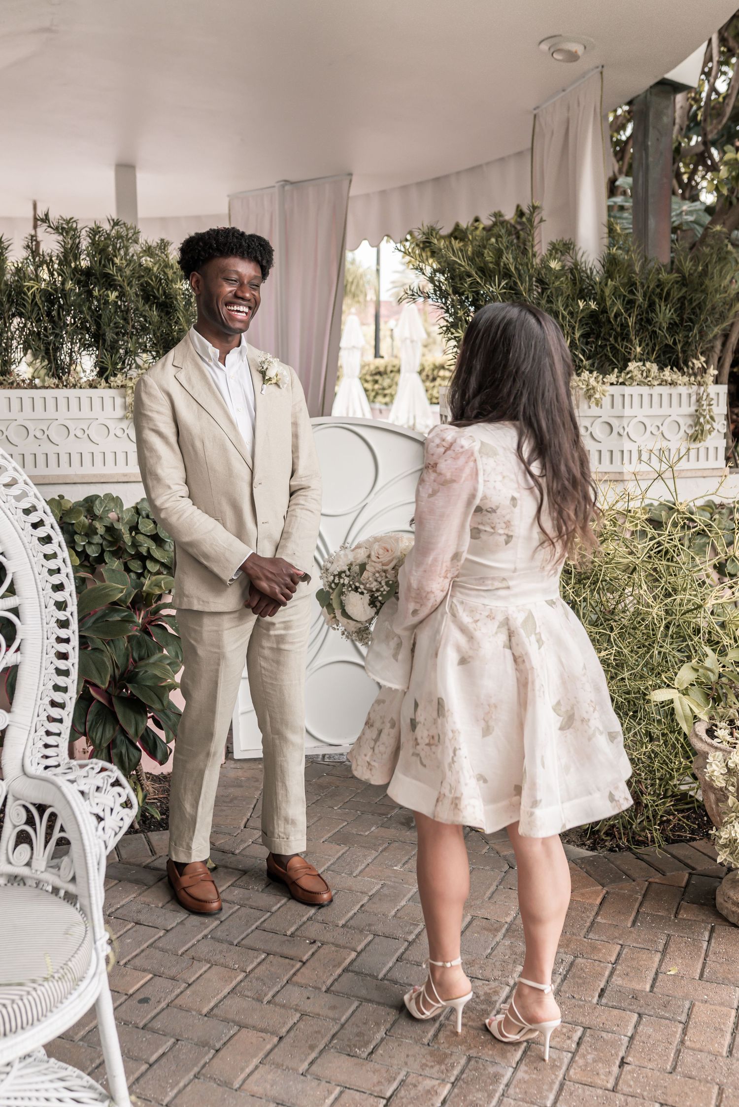 A romantic couple in light-colored outfits poses together on a brick patio with decorative white chairs and lush greenery.