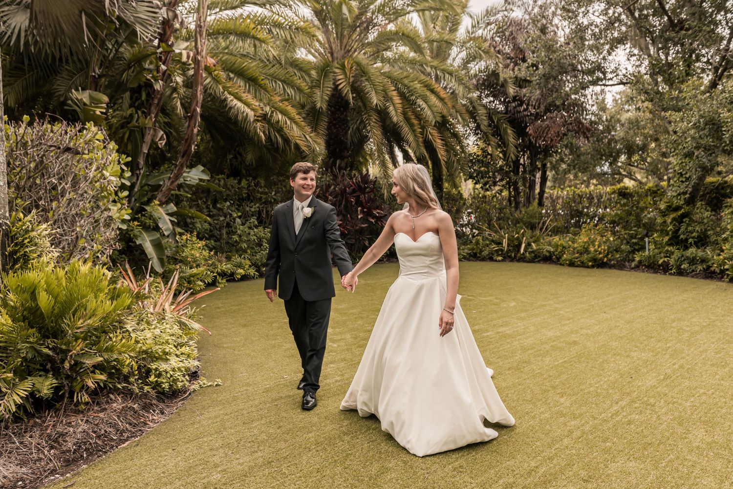 Bride and groom walk hand in hand through a lush tropical garden, bride in elegant white ballgown.