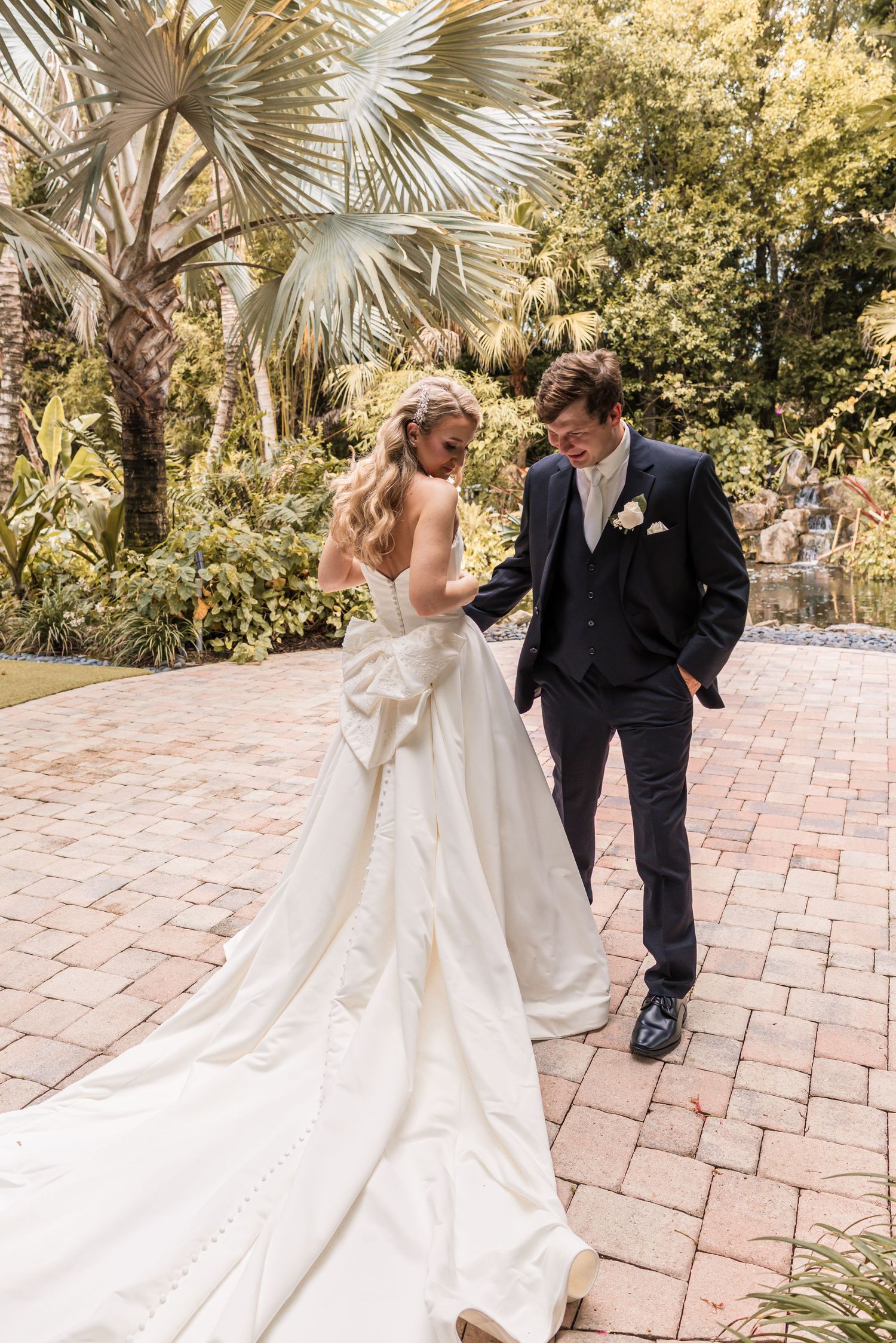 Bride in elegant white gown with train stands beside groom in navy suit on brick patio surrounded by lush tropical greenery.