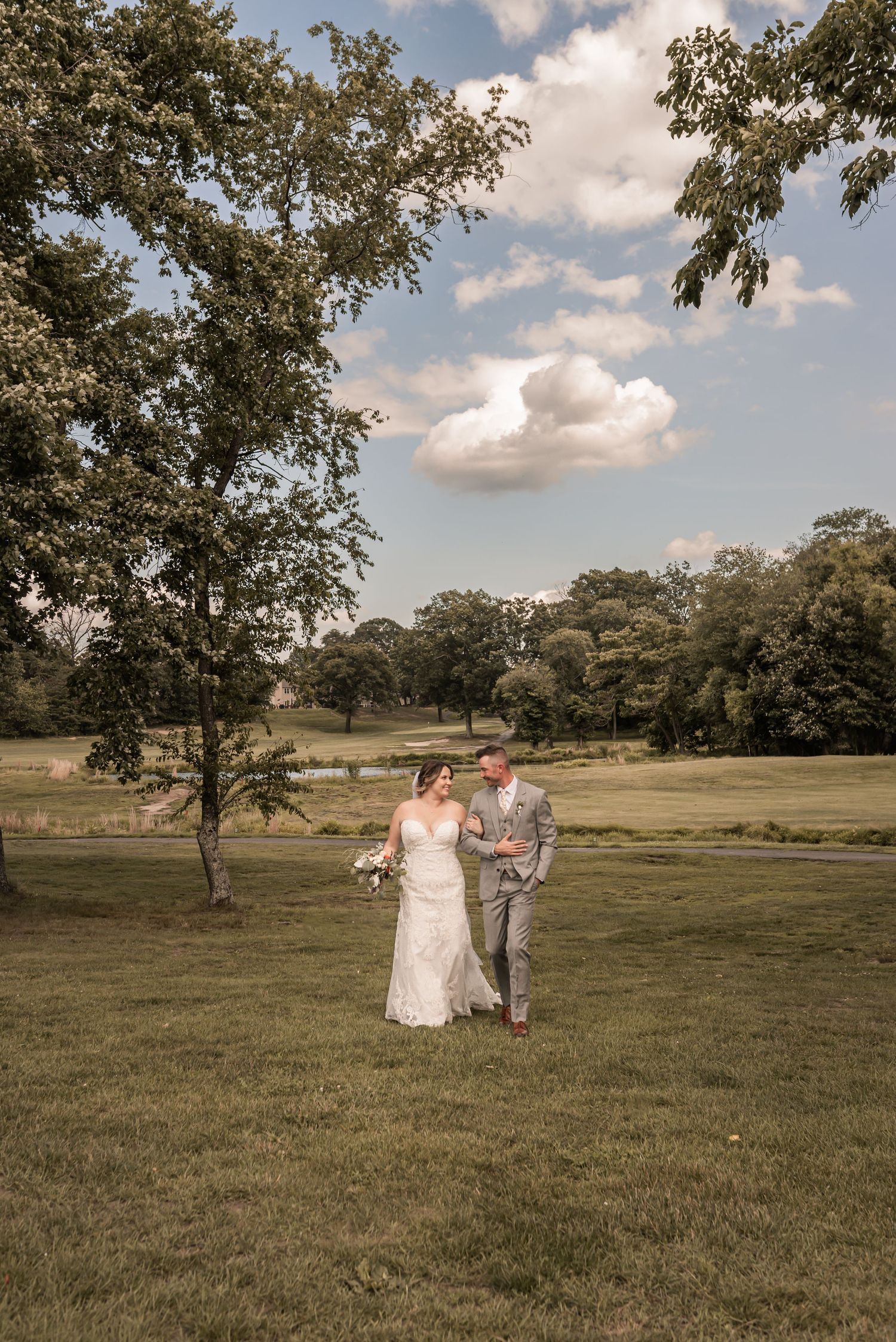 A romantic wedding couple walks together with their dog across a green field on a sunny summer day with puffy clouds overhead.