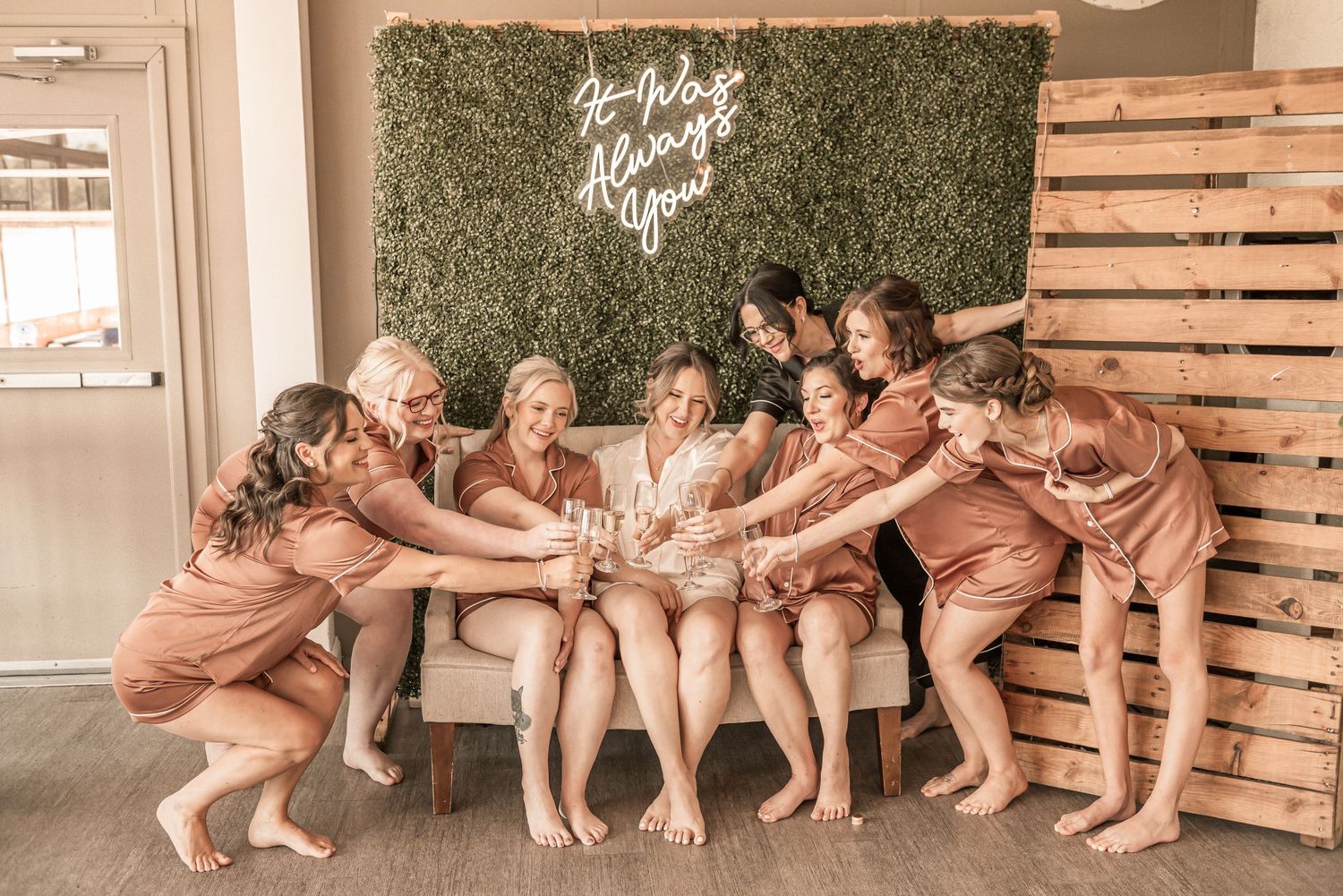 A series of bridesmaids in blush pink dresses pose playfully against a rustic wood wall and greenery backdrop.