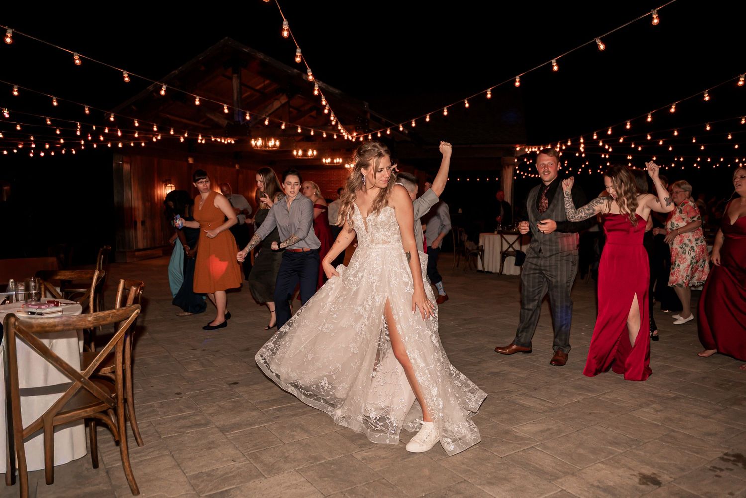 Wedding guests dance together under twinkling string lights at an outdoor evening reception celebration.