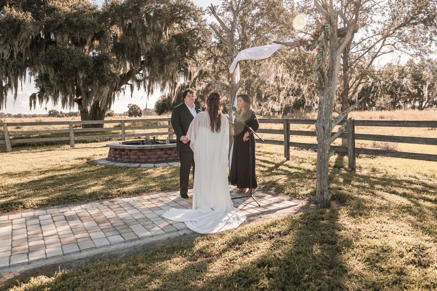 A romantic couple stands beneath Spanish moss draped trees during a golden sunset photo session at a rustic venue.