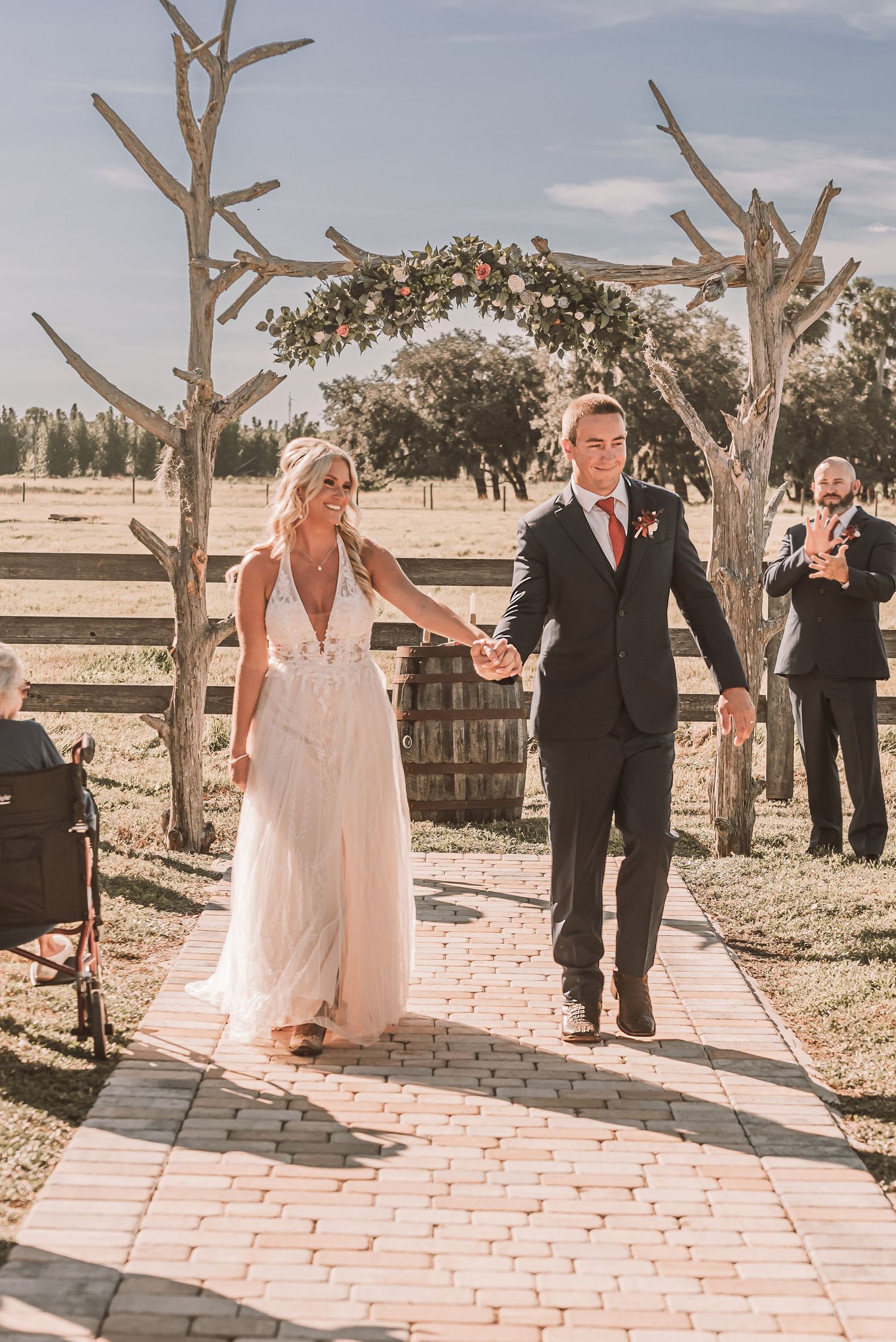 Wedding couple walks down a brick pathway surrounded by desert trees in a rustic outdoor venue setting.