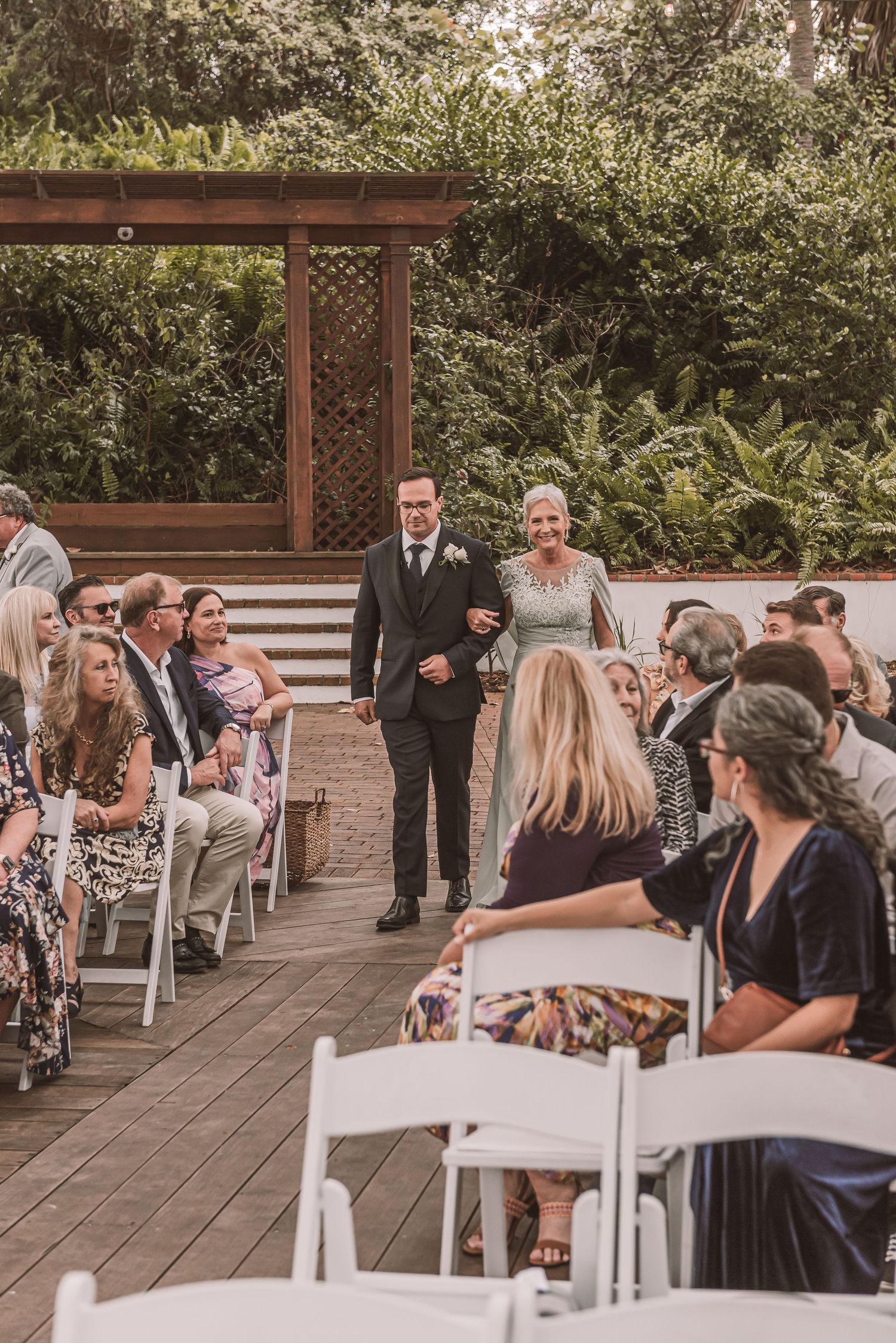 A couple walks down the aisle at an outdoor garden wedding ceremony with guests seated in white chairs on a wooden deck.