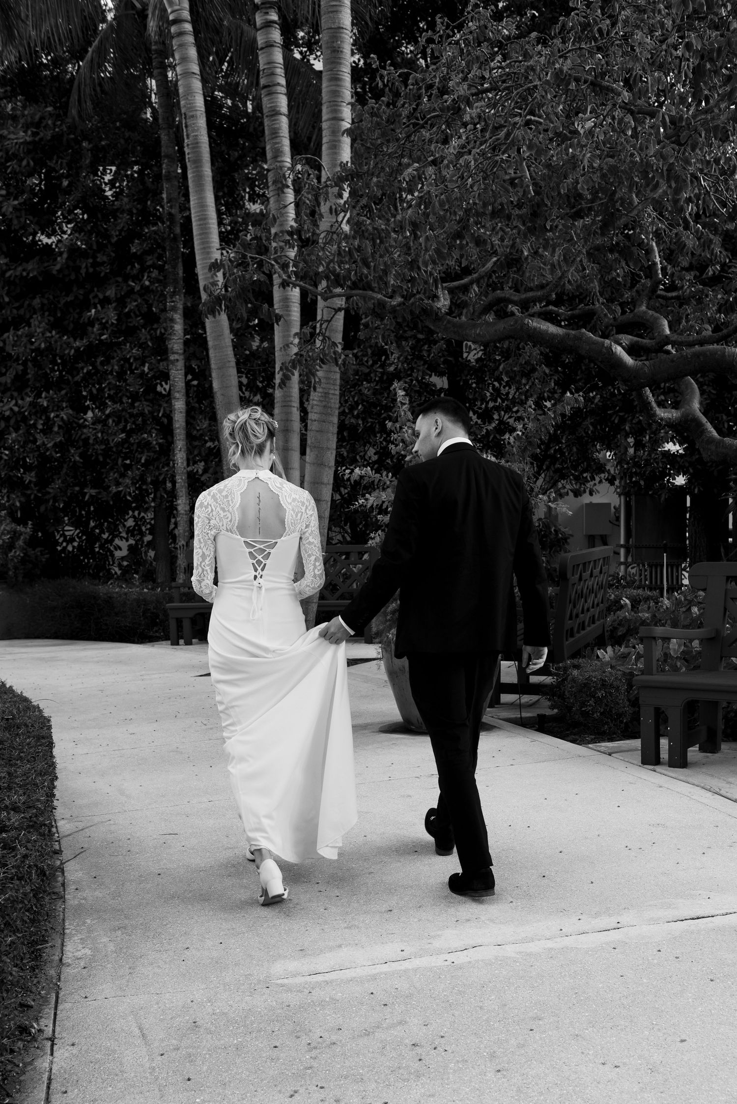 Black and white photo of bride and groom walking hand in hand down a palm tree-lined path on their wedding day.