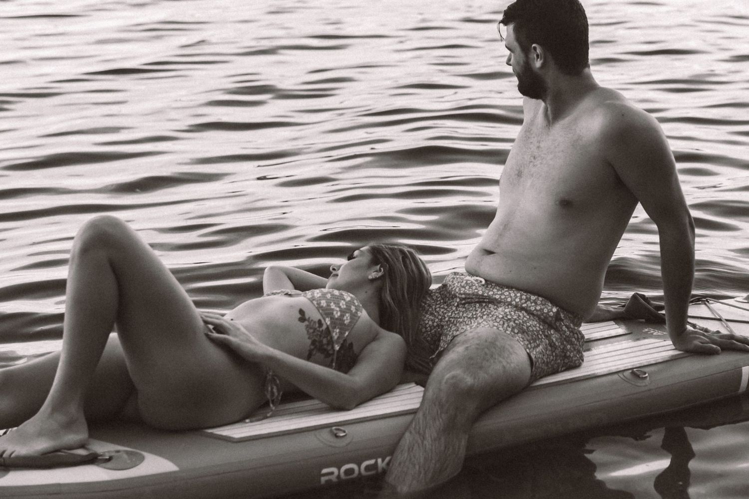 Black and white photo of a couple relaxing on a paddleboard on calm water.