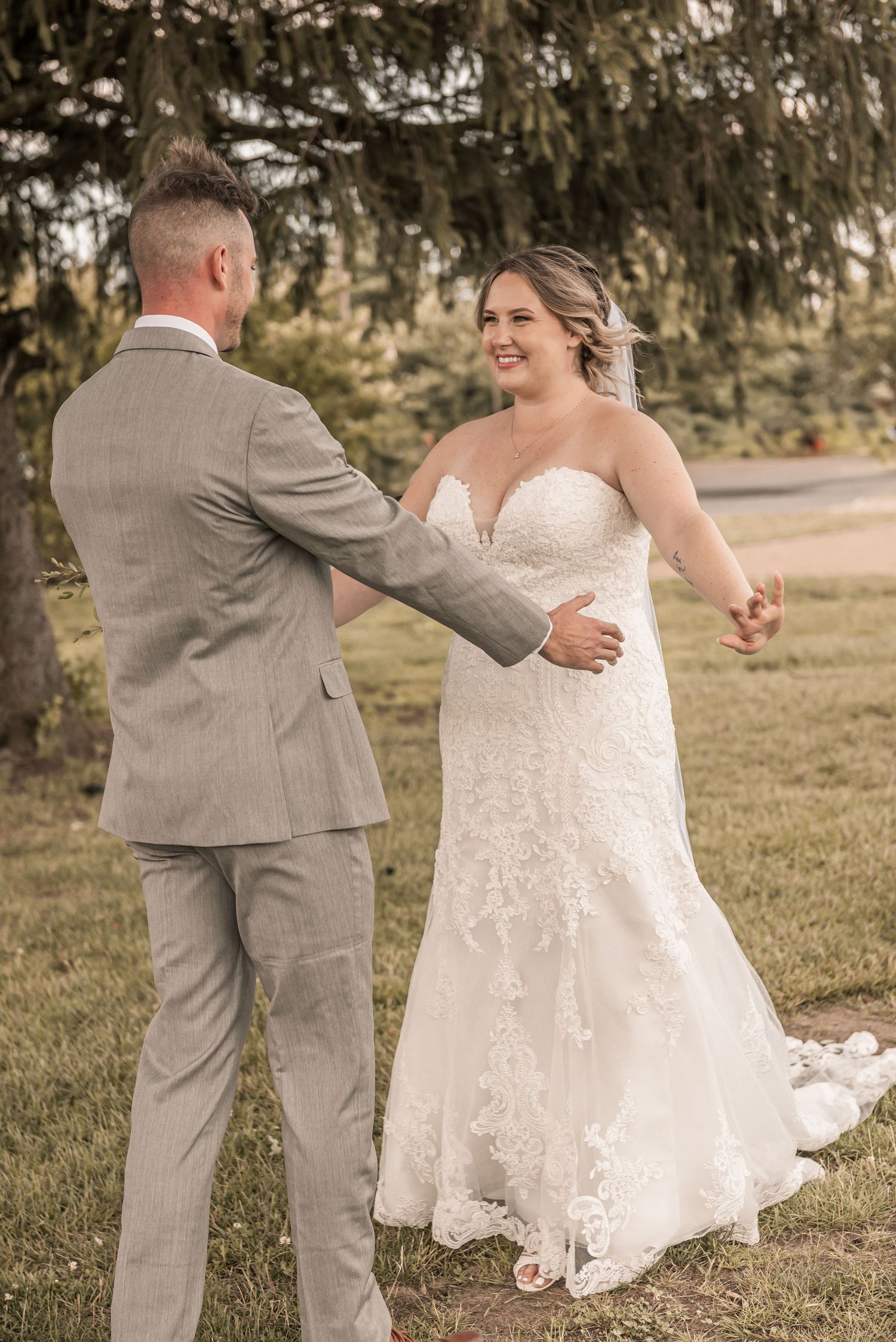 A couple in wedding attire dances together in a grassy field, with the bride's white lace dress flowing gracefully.