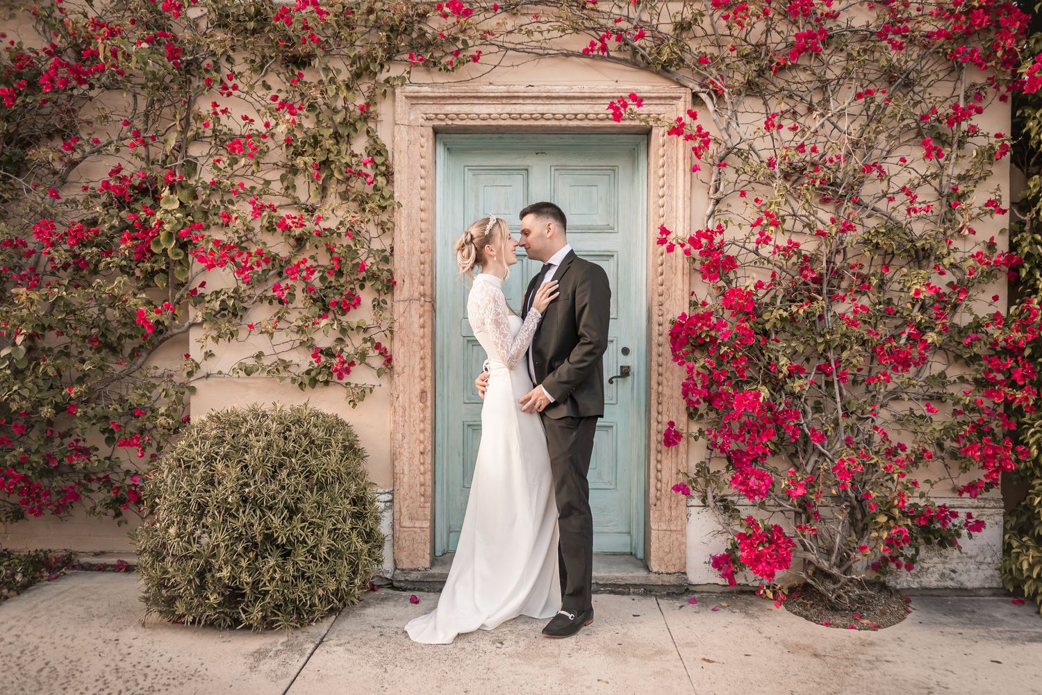 Bride and groom embracing before a teal door surrounded by vibrant red bougainvillea on a beige wall.