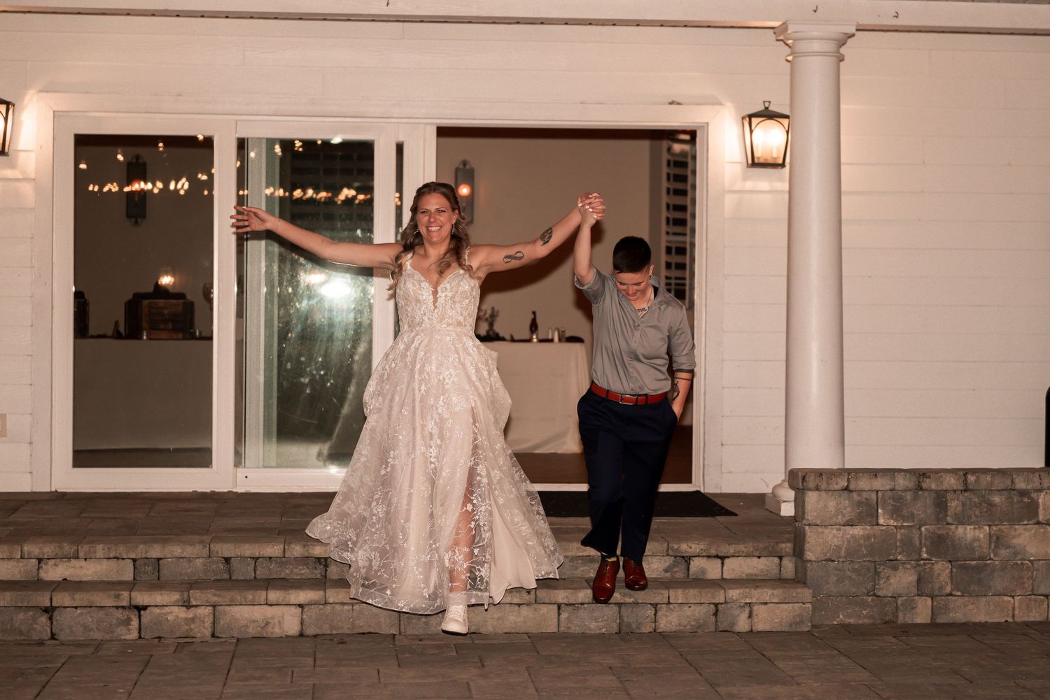 A person in a white lace wedding dress poses playfully on a stone ledge outside a building at dusk.