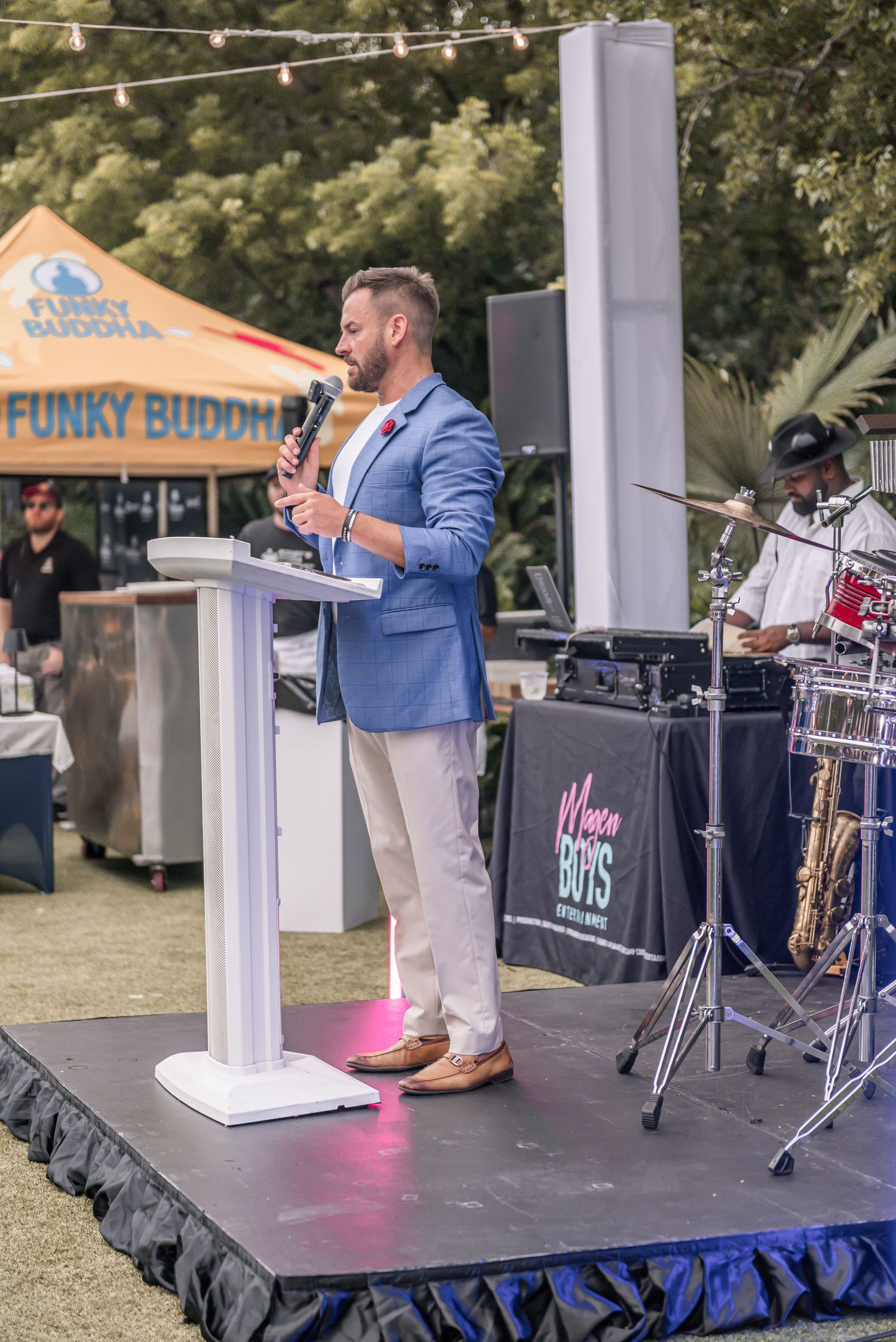 A speaker in a blue blazer and light pants stands at a podium on an outdoor stage with band equipment visible.