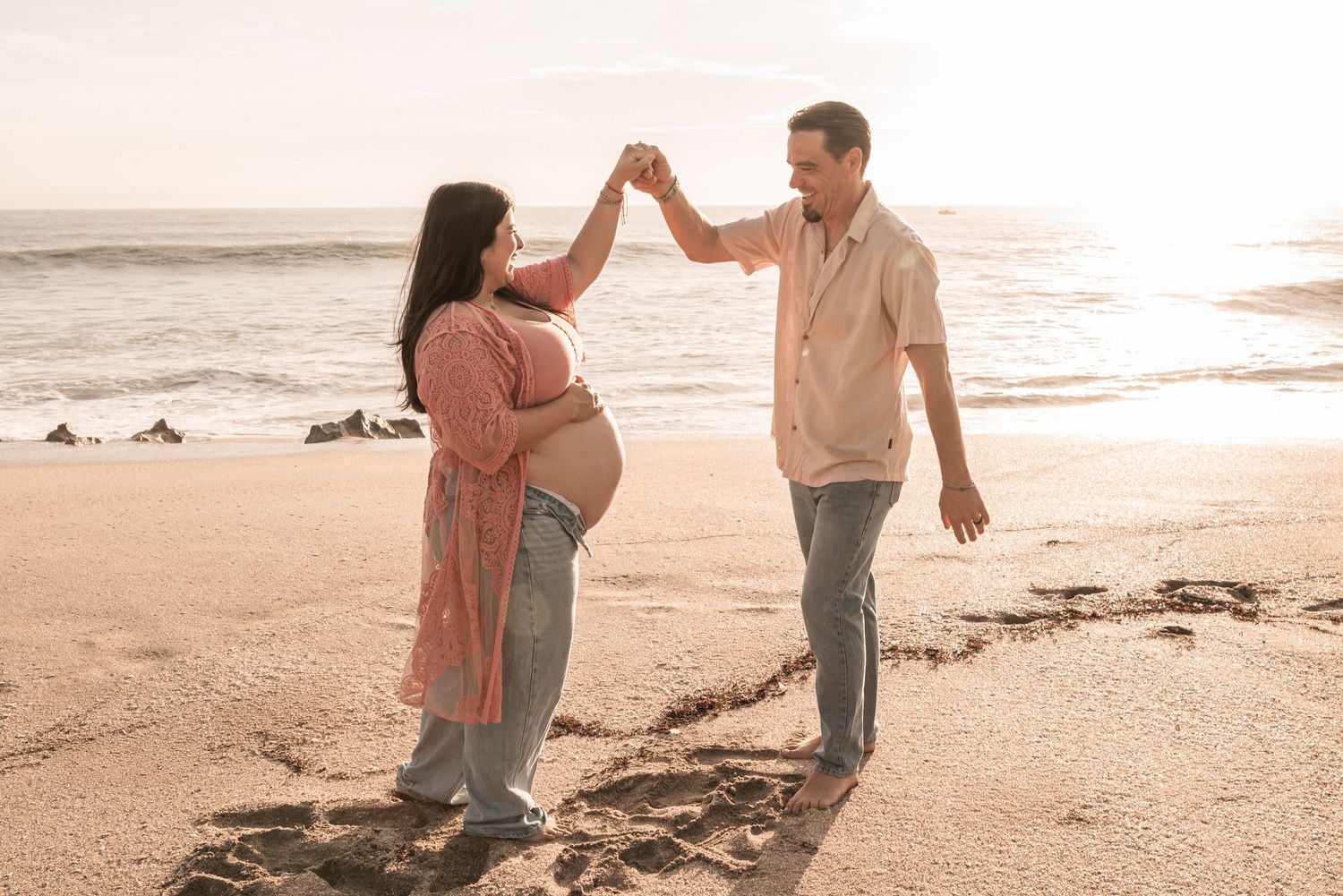 Couple in light colored outfits dancing together playfully on a sandy beach at sunset.