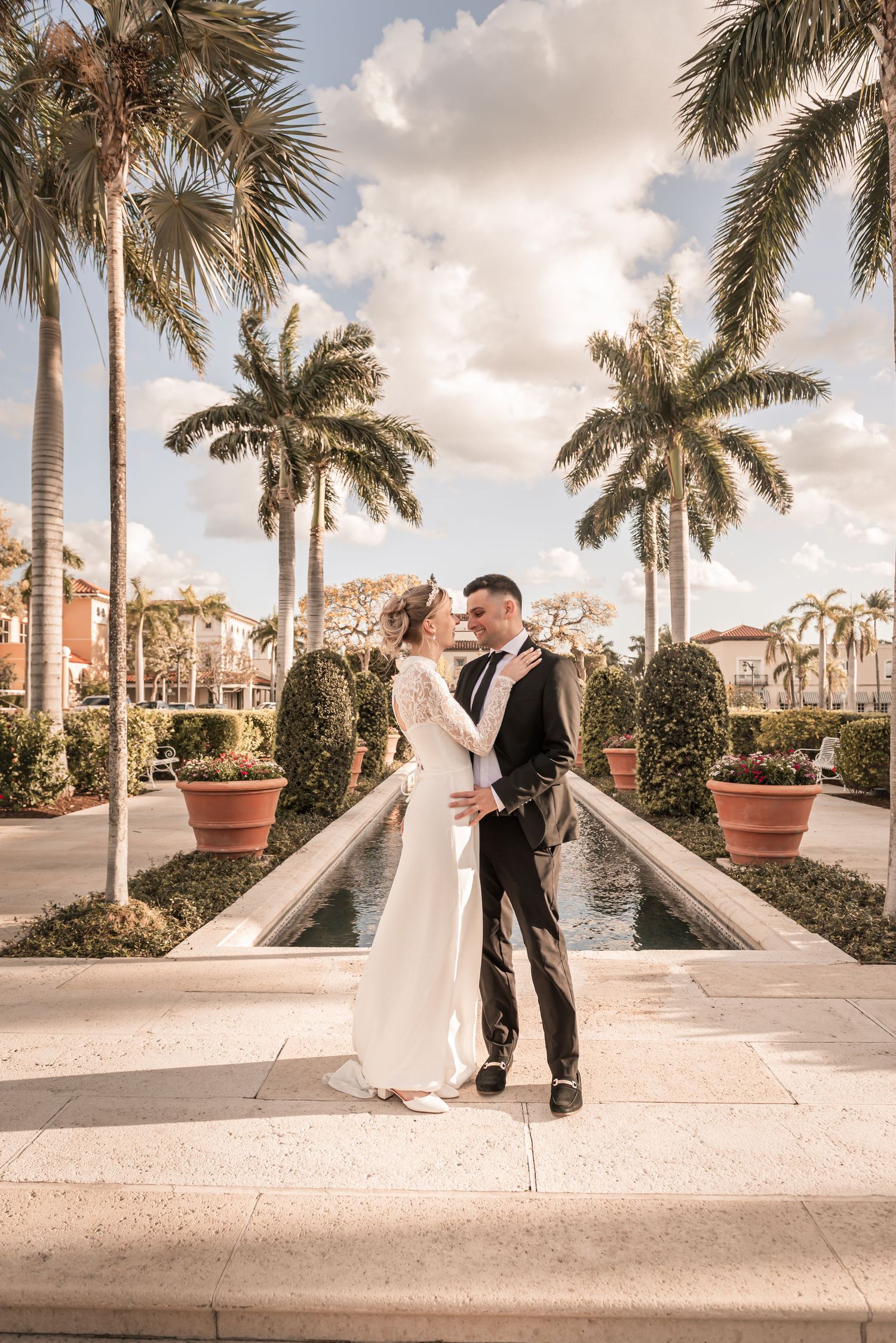 Wedding couple embracing near fountain surrounded by tall palm trees at tropical venue under cloudy sky.