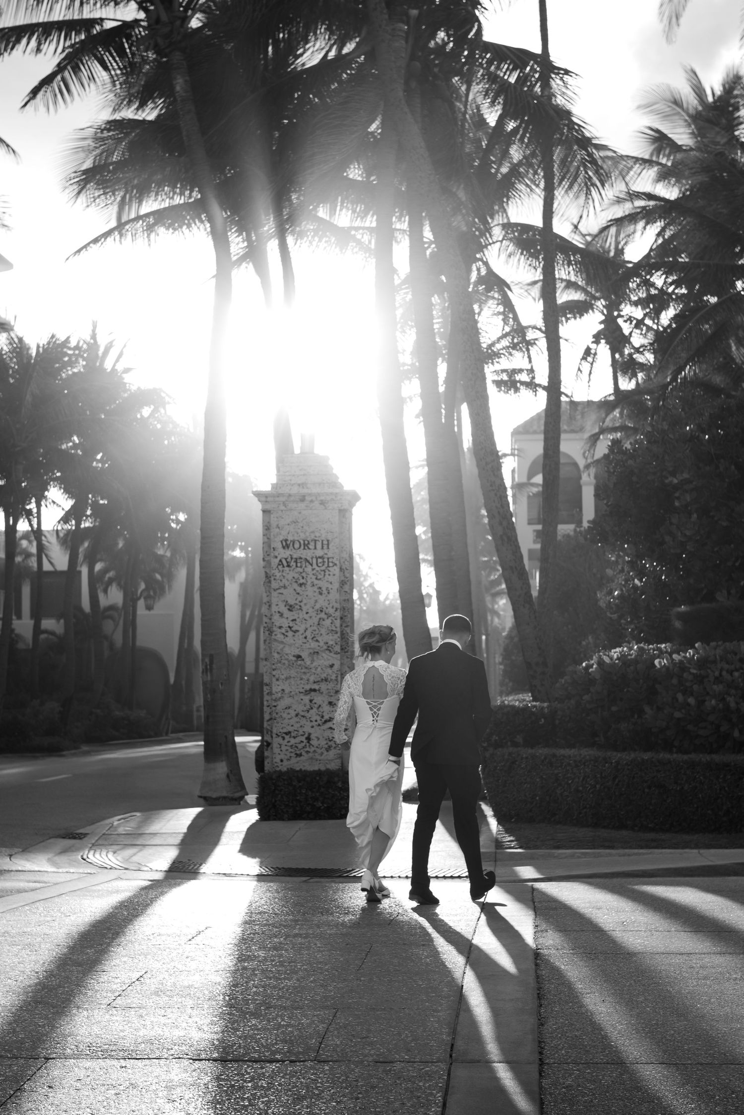 Black and white photo of a bride and groom walking past palm trees in bright sunlight on their wedding day.