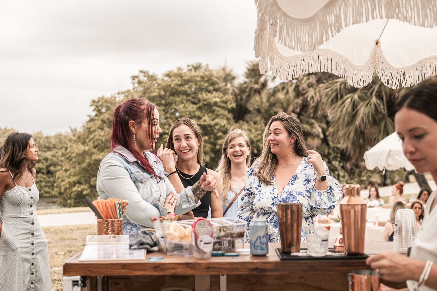 A group of friends gather around a rustic outdoor table with parasols enjoying drinks and laughter at a summer party.