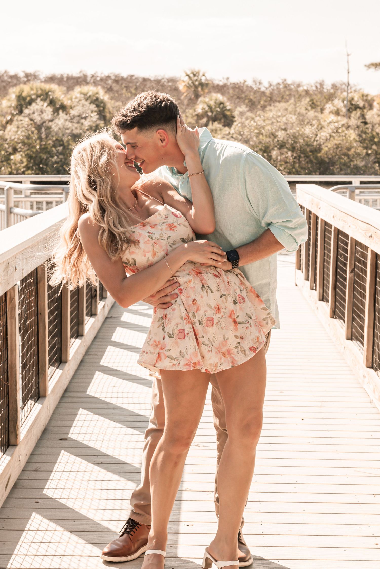 Couple sharing a romantic kiss on a sunny wooden boardwalk, woman in floral dress, man in light blue shirt.