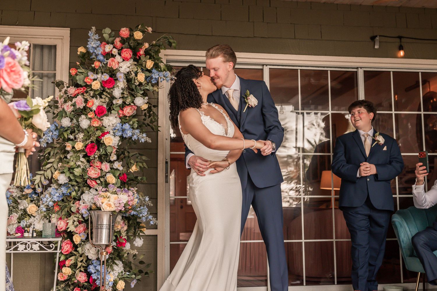 Bride and groom share first dance beside colorful floral arrangement at wedding reception.