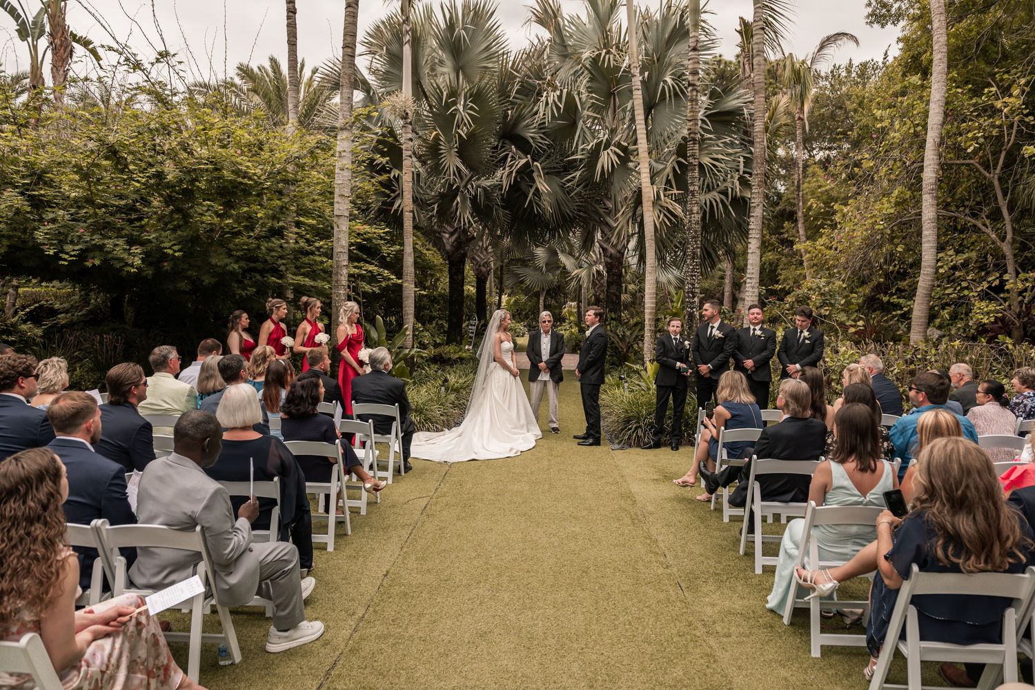 Outdoor wedding ceremony on green lawn with bride and groom at altar, guests seated in white chairs, surrounded by tall palm trees.
