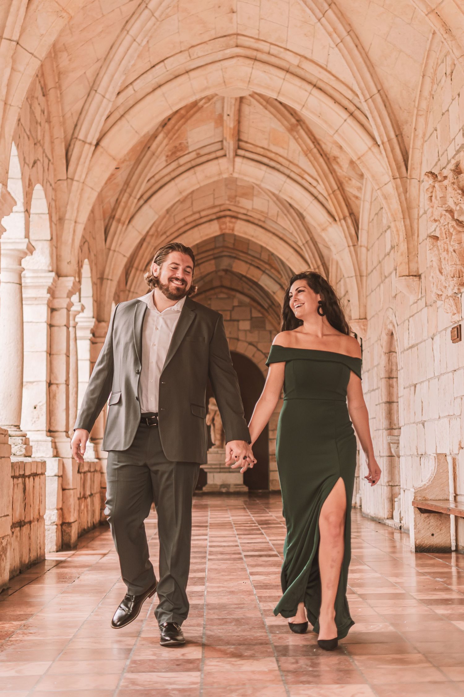Couple in formal attire poses playfully in a historic stone archway corridor with repeating arched columns.