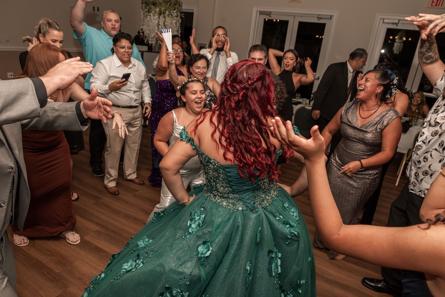 Young woman in a green quinceañera gown dancing joyfully surrounded by celebrating guests at a party.