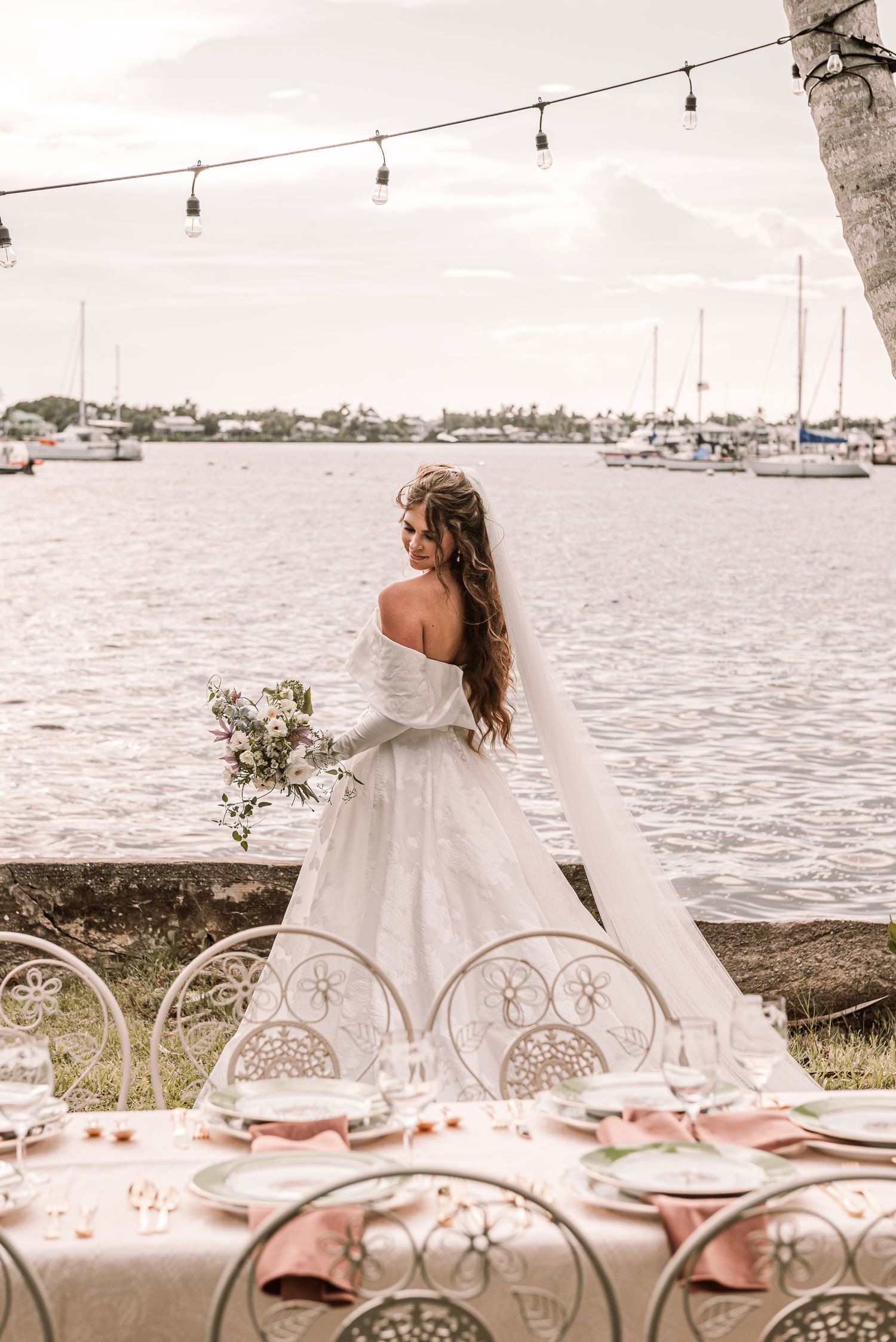 An elegant waterfront wedding reception setup with string lights, white tablecloths and vintage chairs overlooking the marina.