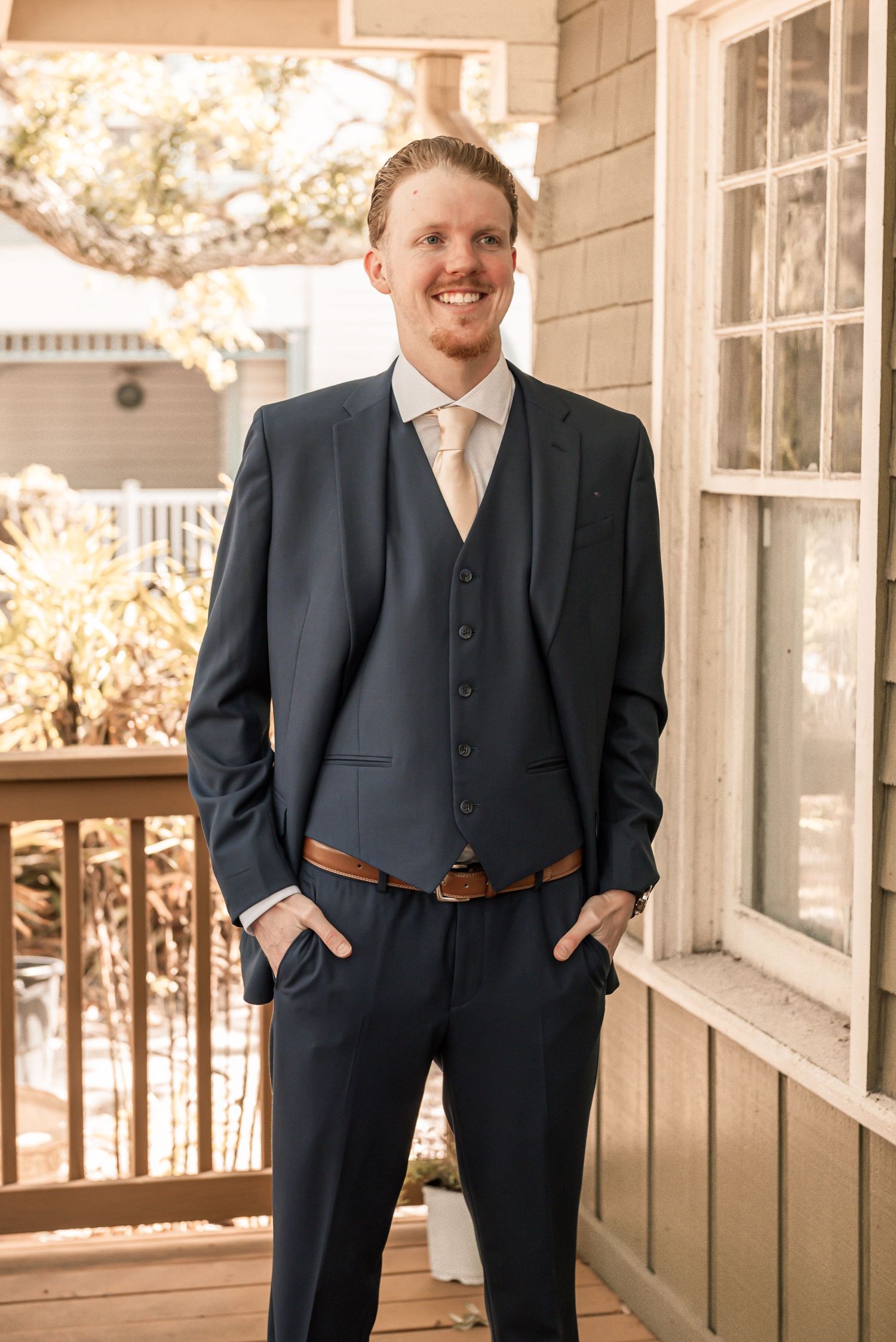 Smiling groom in navy three-piece suit with tan tie and brown belt posing on a wooden porch.