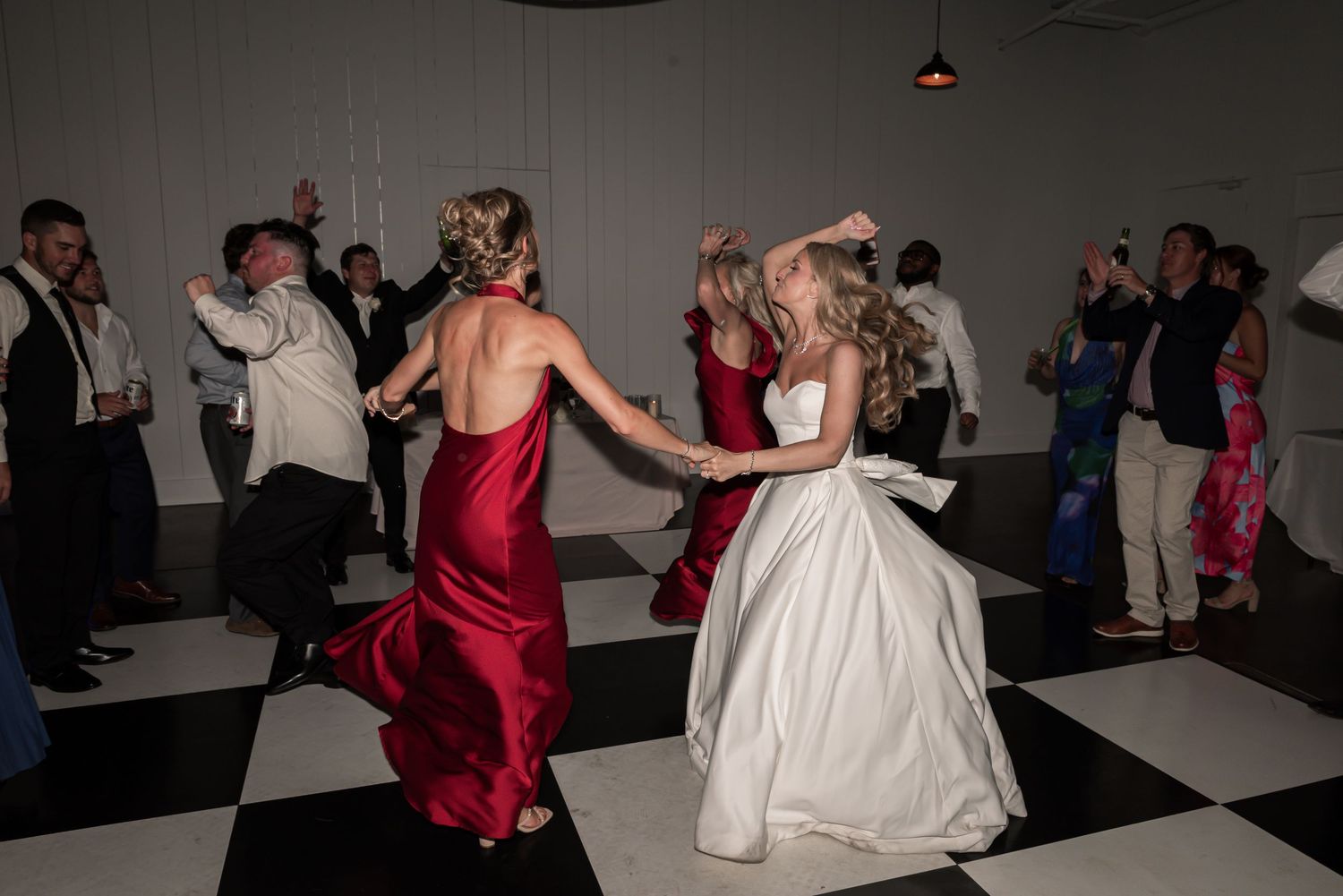 Bride in white gown dancing with guest in red dress on checkered dance floor at wedding reception.