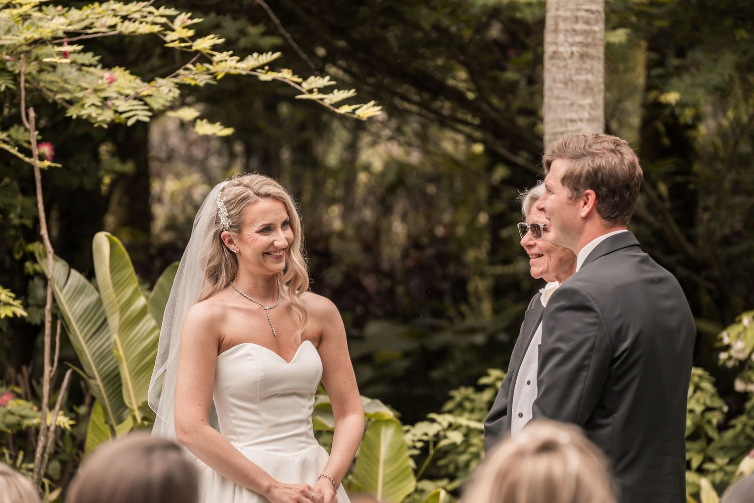 Bride in white gown and veil smiling at groom during outdoor wedding ceremony surrounded by lush tropical greenery.