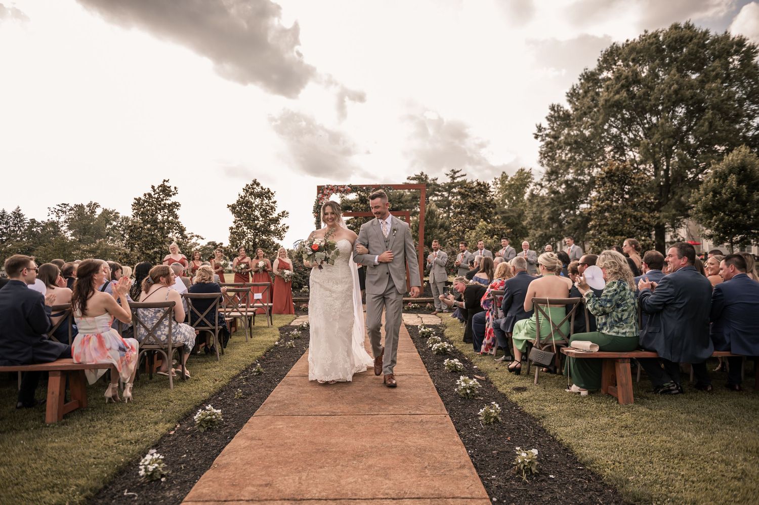 A rustic outdoor wedding ceremony with guests seated on wooden benches along a brick aisle under cloudy skies.
