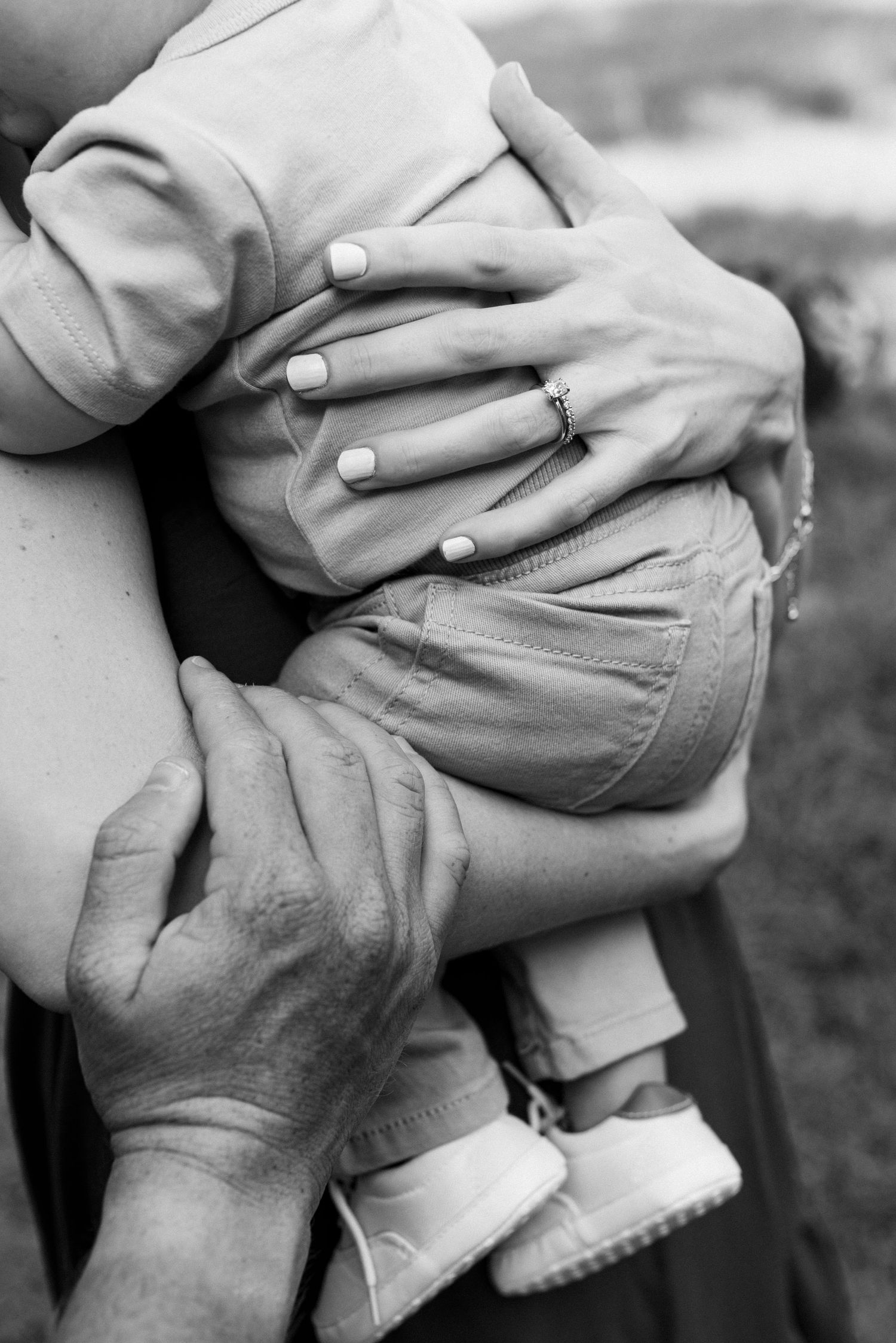 Black and white close-up of hands holding a small child wearing shoes, with a diamond ring visible on one hand.