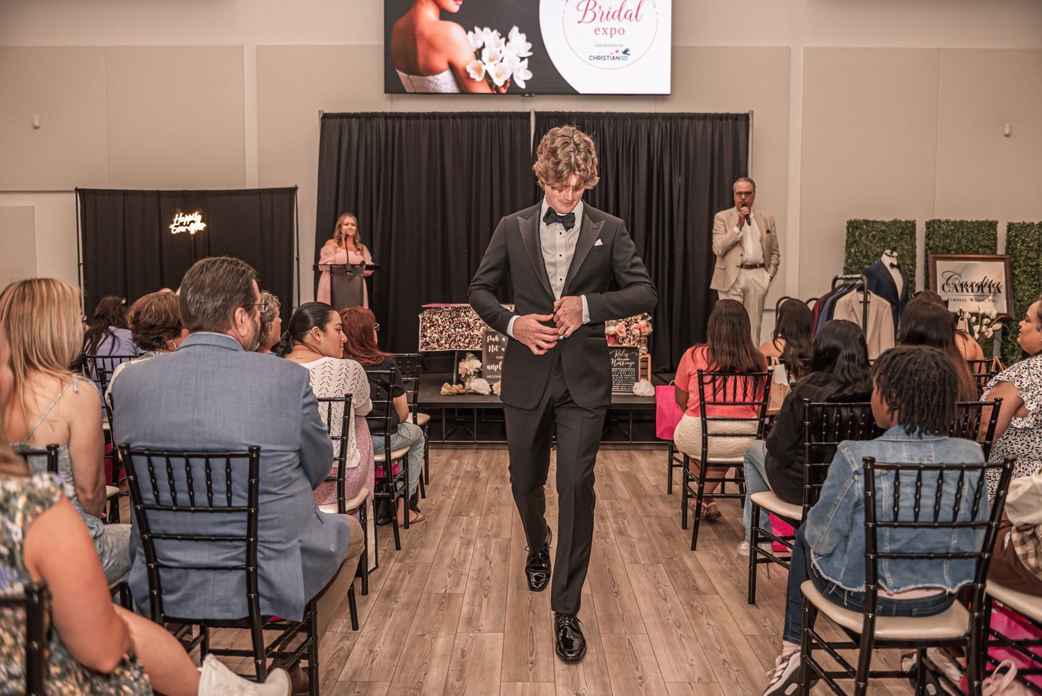 A person in a black suit walks down a wedding venue aisle lined with chairs and guests during an indoor ceremony.