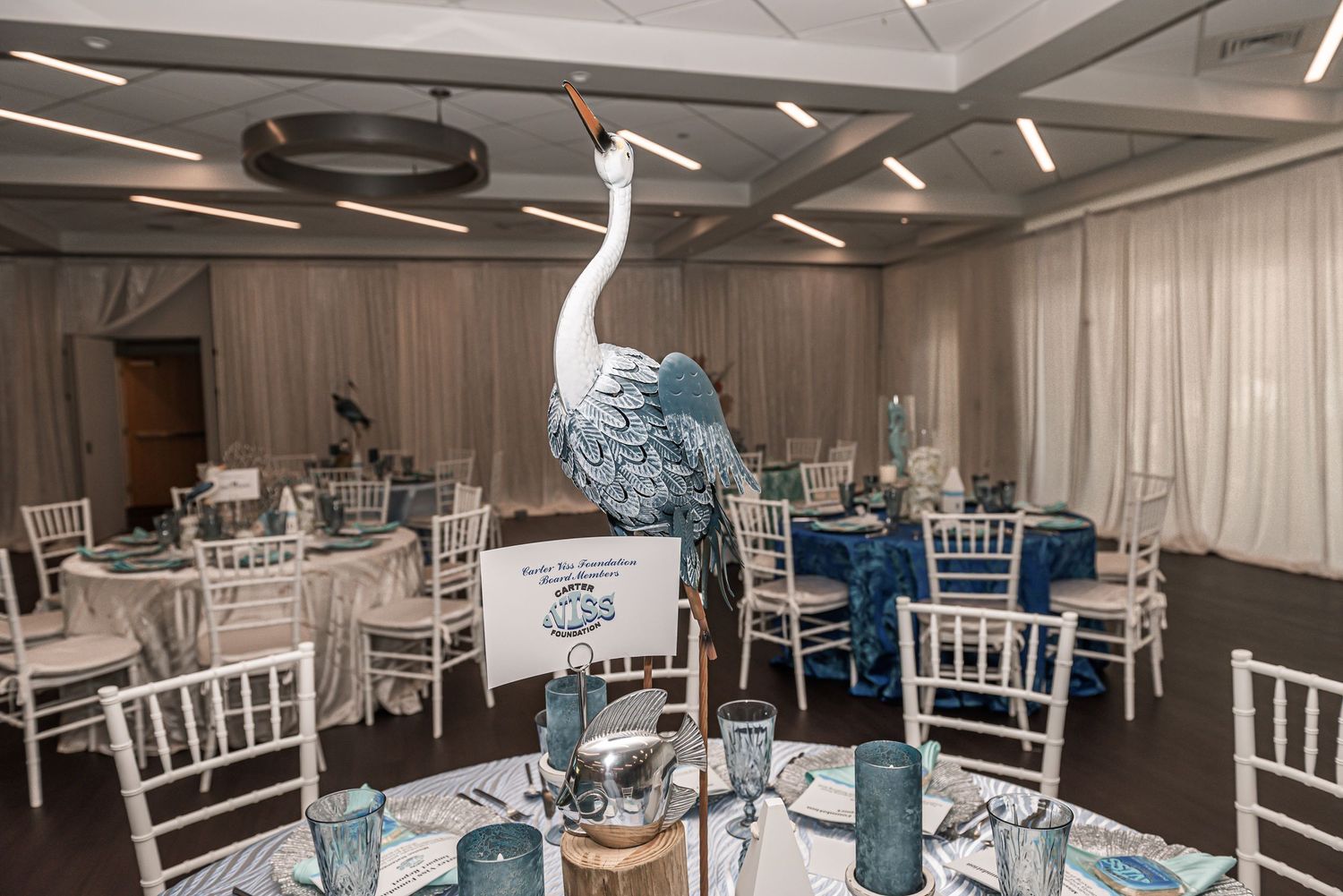 Elegant wedding reception table setting with white chairs and tall peacock centerpiece in a modern banquet hall.