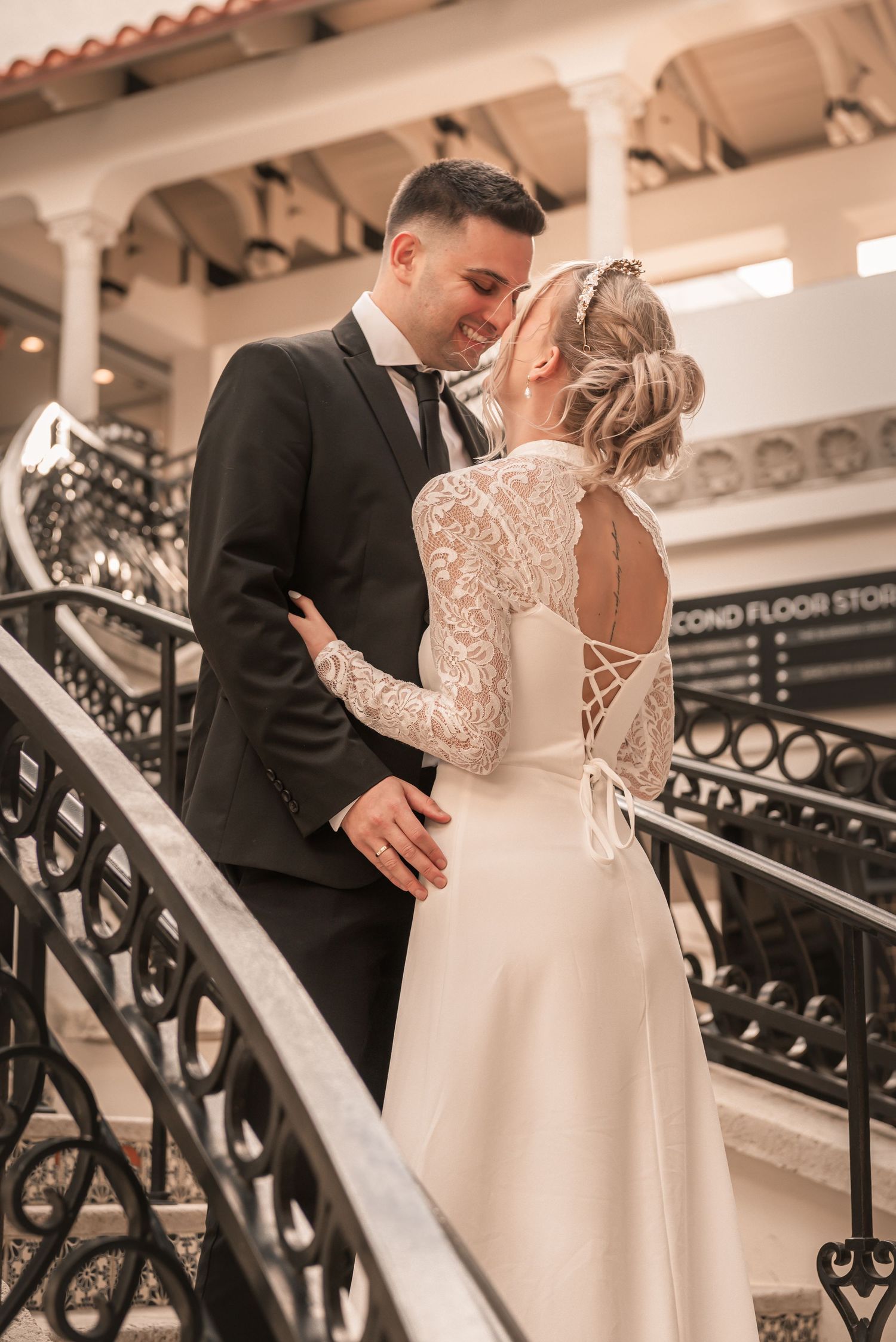 Bride and groom sharing a romantic moment on ornate staircase, bride in lace back wedding gown, groom in black suit.