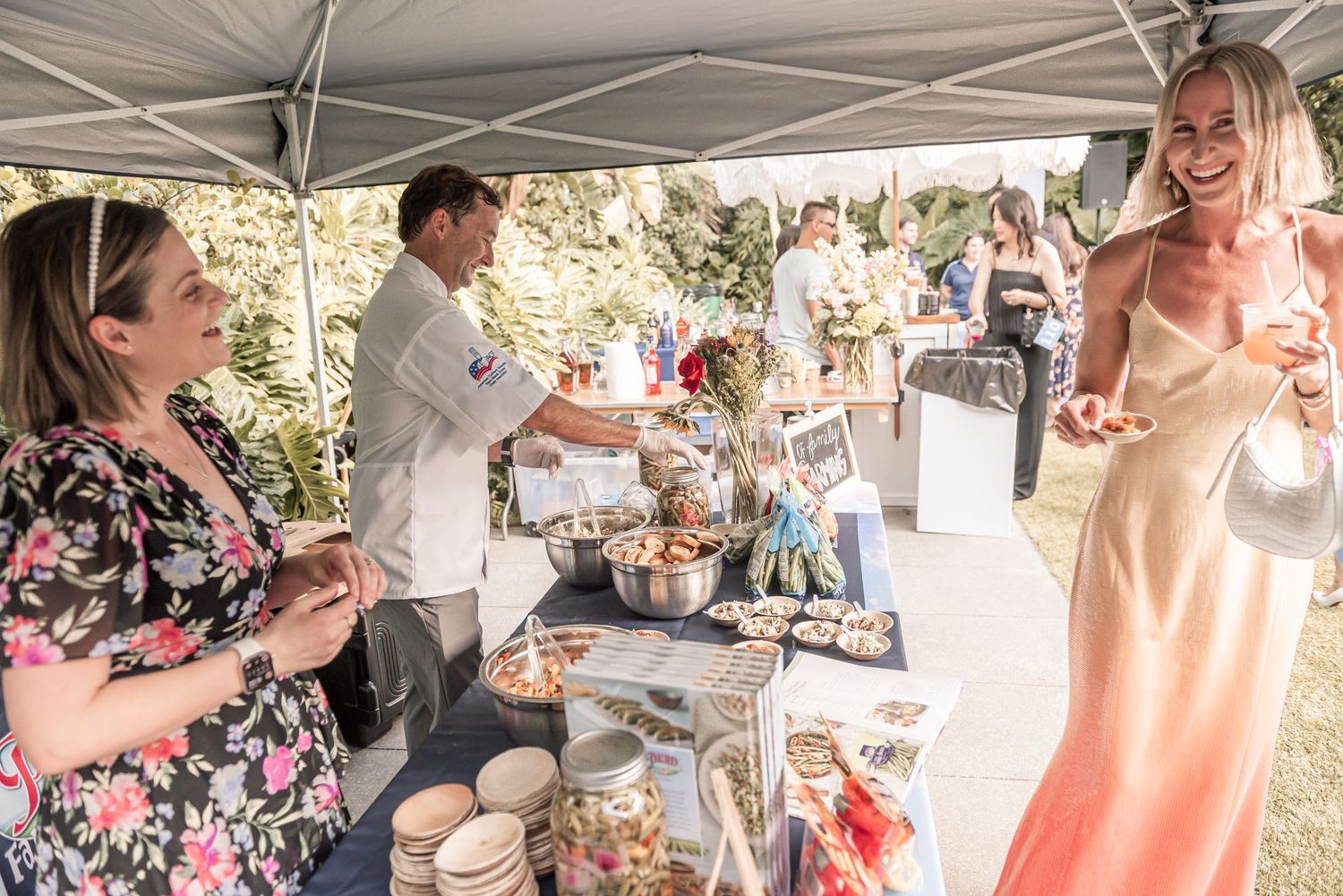 People gather under a white tent at an outdoor event serving food and drinks on a sunny day.