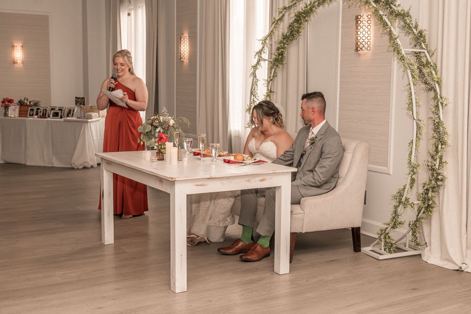 Wedding reception setup featuring floral arch, white table, and elegant decor in light-filled venue.