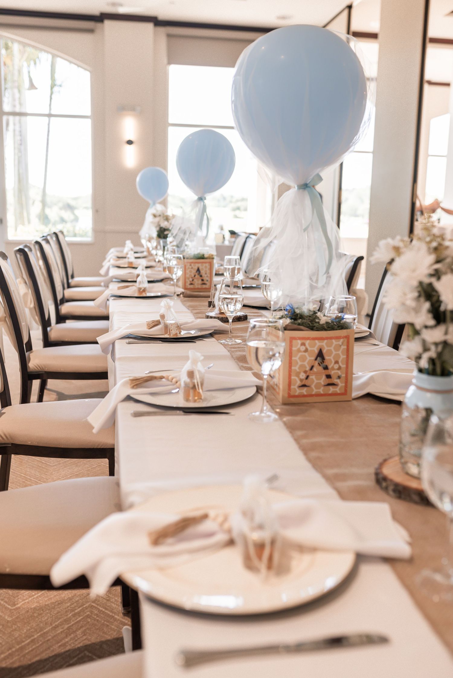 Baby shower table setting with blue balloons, white plates, and wooden alphabet blocks in a bright reception room.