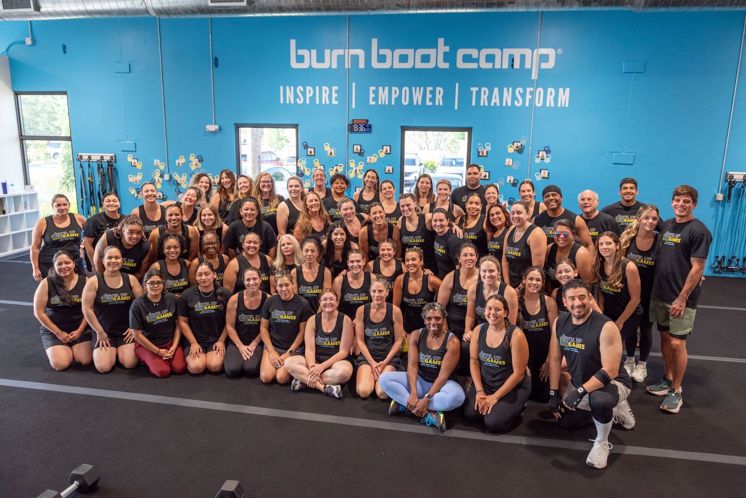 Large group of Burn Boot Camp members and trainers posing together in front of teal Inspire Empower Transform wall.