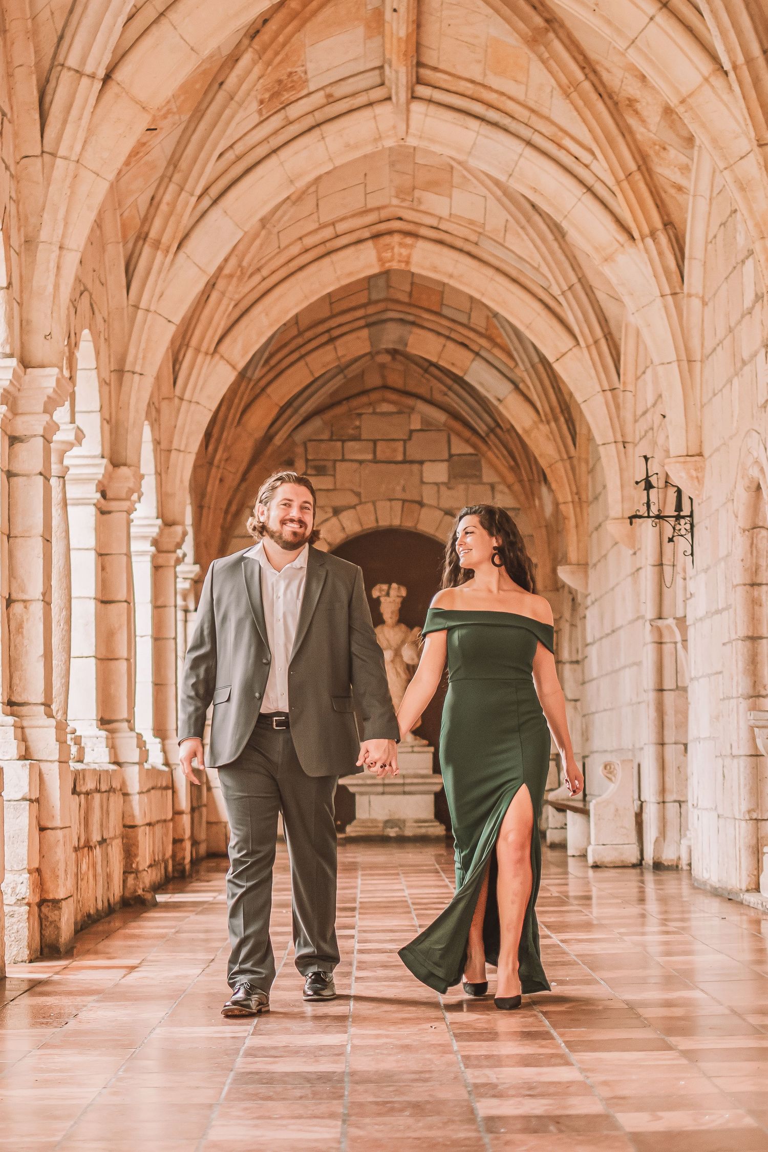 Couple in formal attire poses playfully in a grand stone archway corridor with gothic architecture.