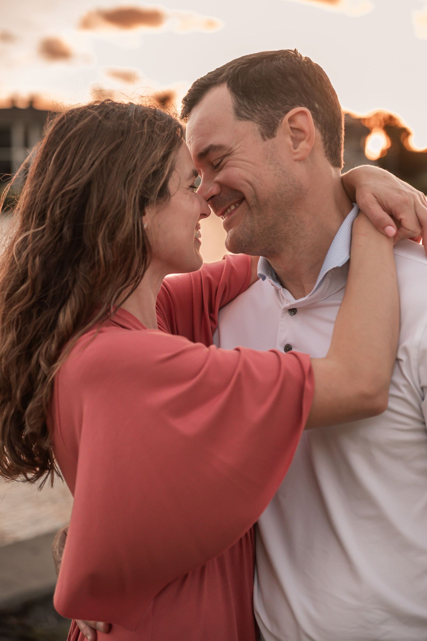 Romantic couple embraces and shares intimate moment at sunset while wearing coral and white clothing.