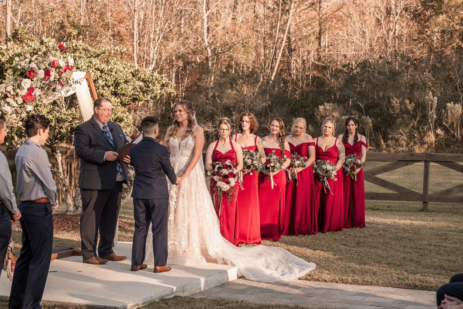 A wedding ceremony takes place outdoors during golden hour with bridesmaids in red dresses standing in a row.