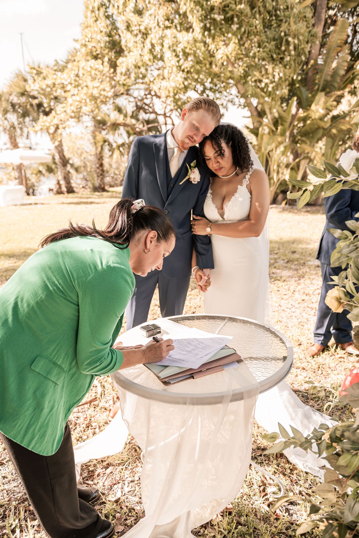 A bride and groom smile as a woman in green signs their wedding register at an outdoor ceremony.