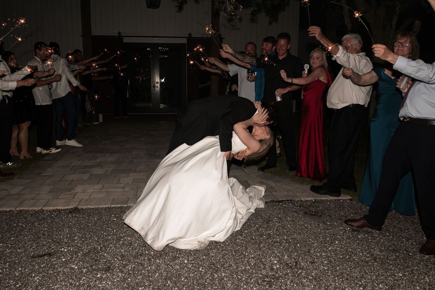 Bride and groom share romantic dip kiss surrounded by guests holding sparklers at outdoor wedding reception.