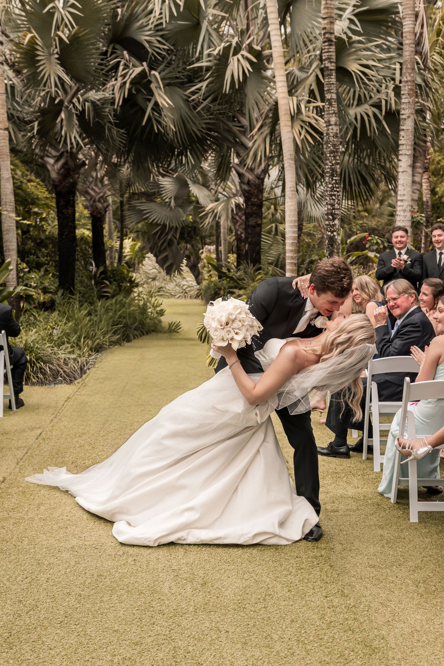Groom dips bride for a romantic kiss at outdoor wedding ceremony surrounded by tropical palm trees.