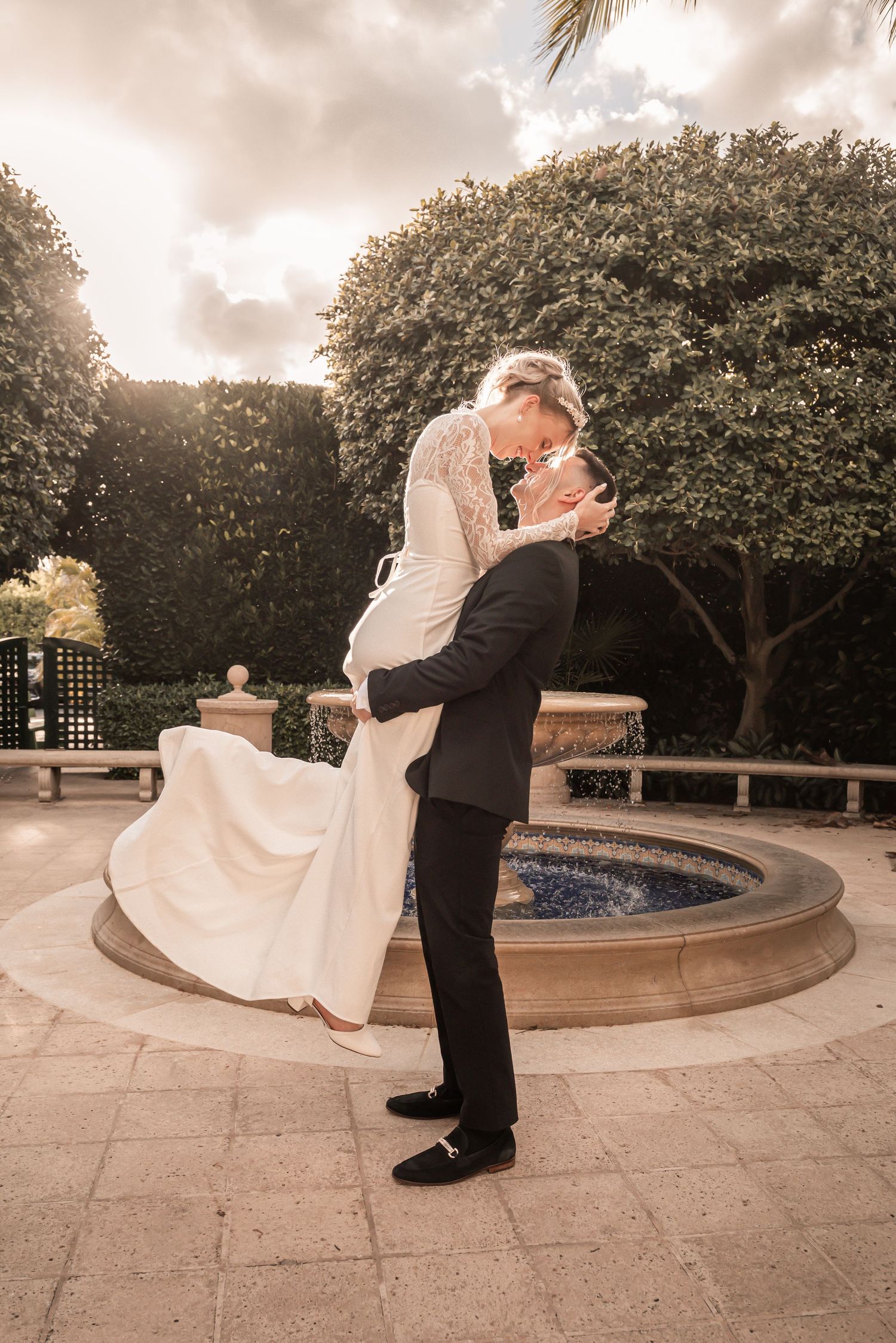 Groom lifting bride in white gown near fountain, sharing romantic kiss outdoors at elegant garden wedding venue.