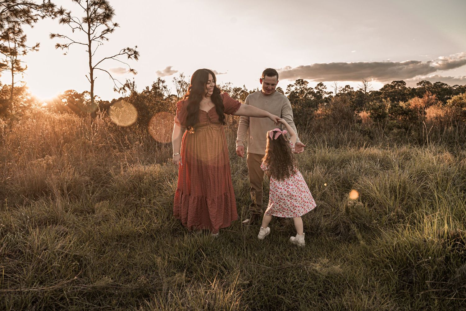 A warm sunset silhouettes a family together in a grassy field with tall pine trees in the background.