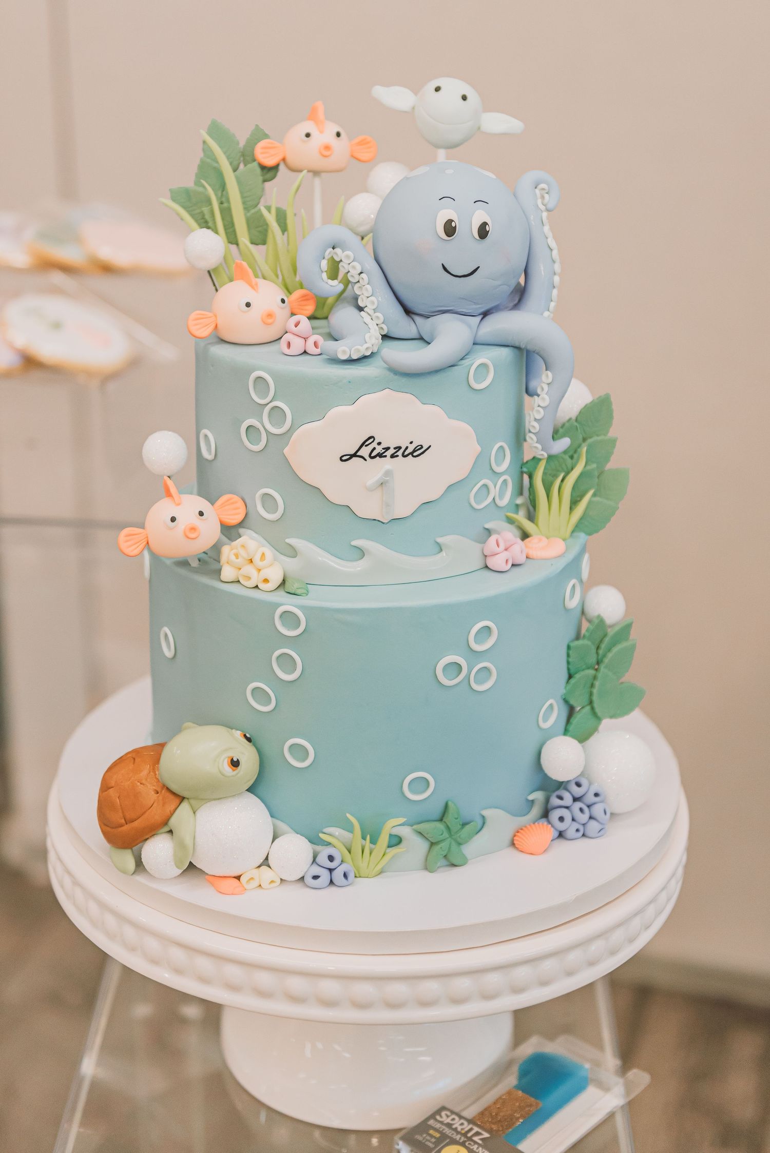 A two-tier ocean themed baby shower cake decorated with fondant sea creatures, bubbles and seaweed in soft blue tones.