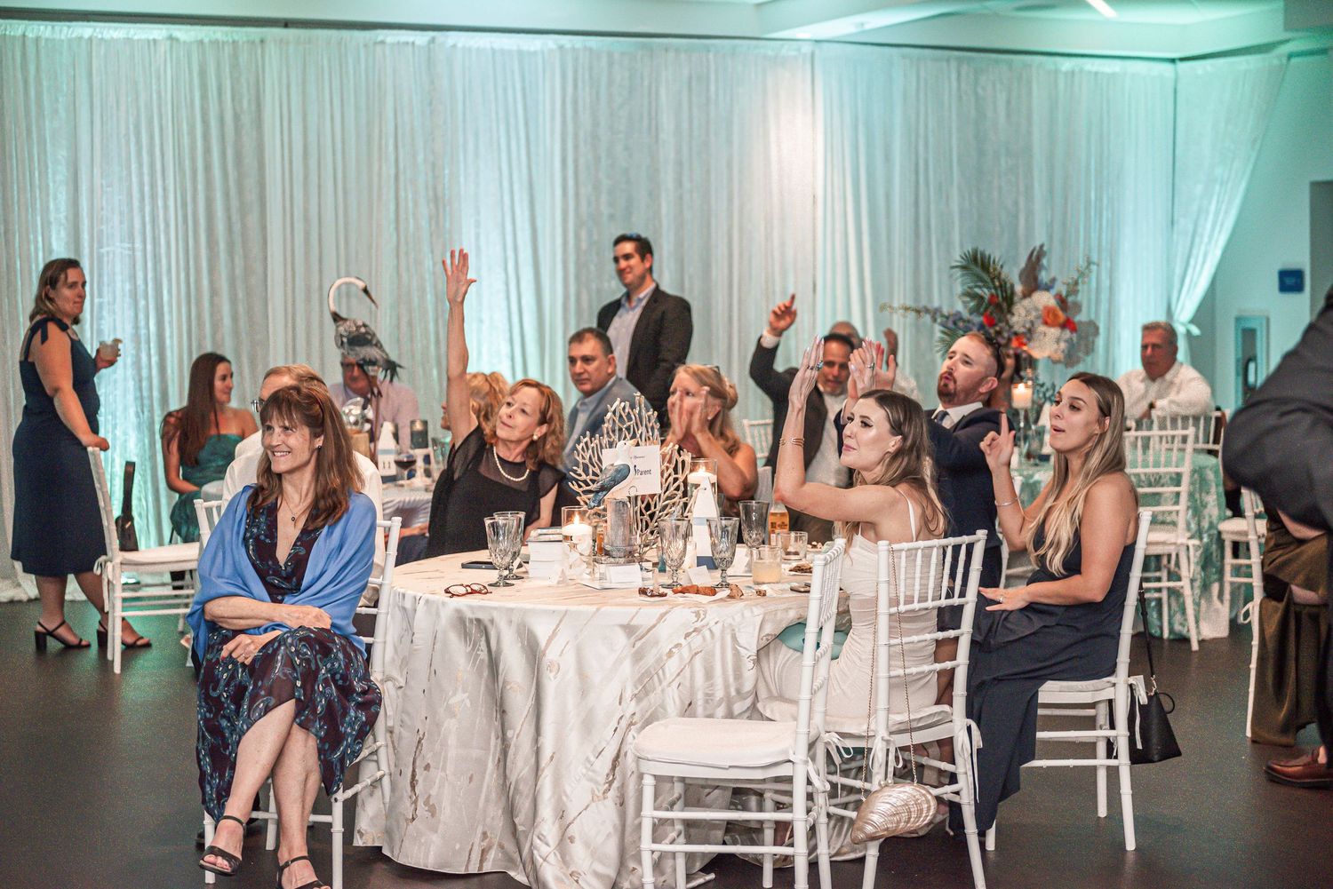 Wedding guests seated at tables raise their hands in celebration during an elegant reception with teal lighting and white decor.